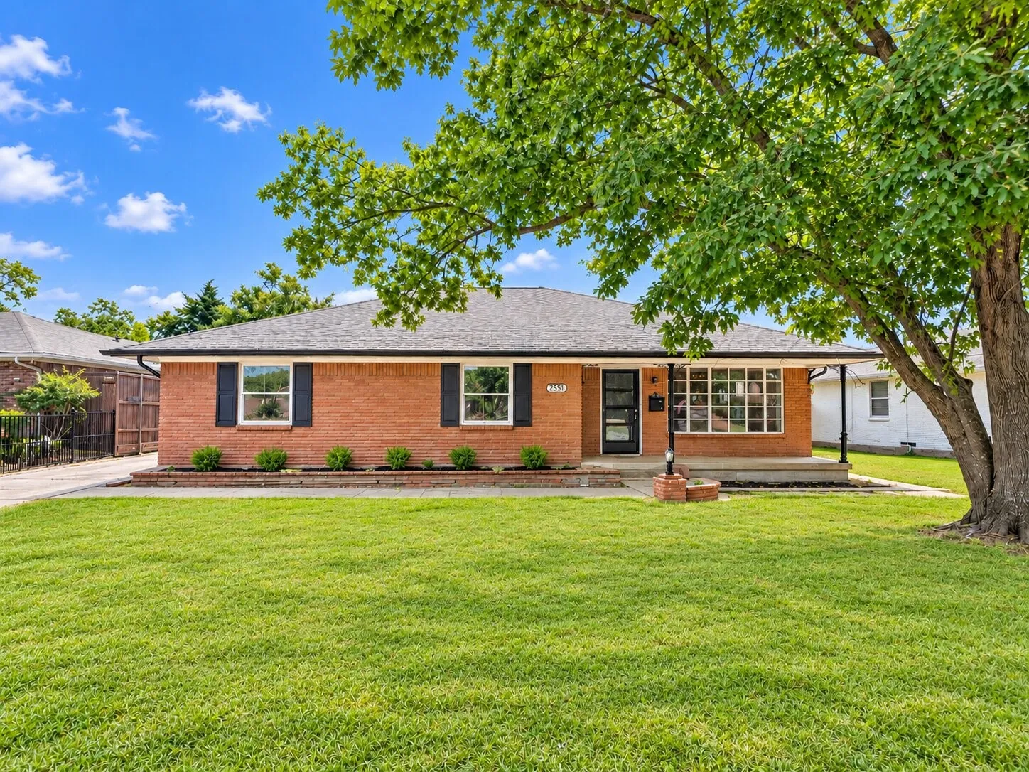 Single-story brick exterior featuring a hip roof, expansive front lawn, shuttered windows, and a covered porch with a brick-surround support pillar
