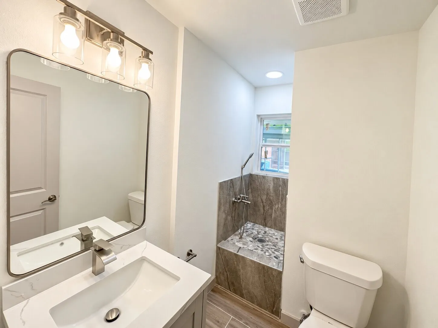 Bathroom featuring a vanity with a white countertop and integrated sink, a rectangular mirror with rounded edges, a three-light fixture with cylindrical glass shades, and a shower area finished with stone-textured wall tile and stone pebble flooring