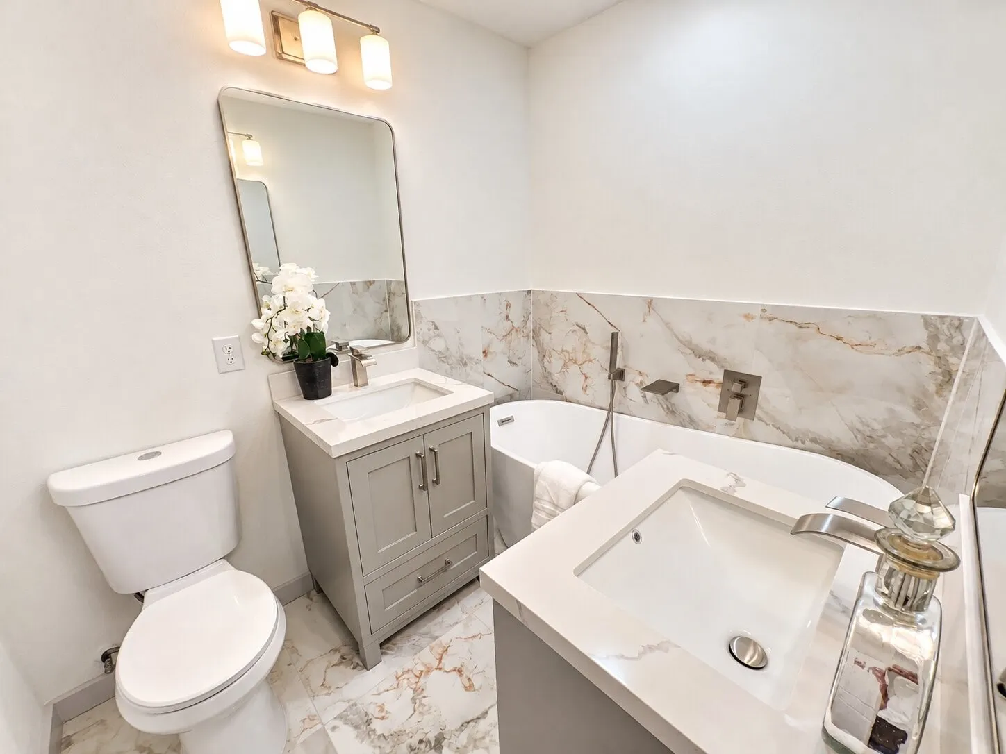 Bathroom featuring dual grey vanities with solid-surface countertops, soaking tub with wall-mounted hardware, floor-to-ceiling stone-patterned tile wall cladding, and oversized polished stone-patterned floor tiling