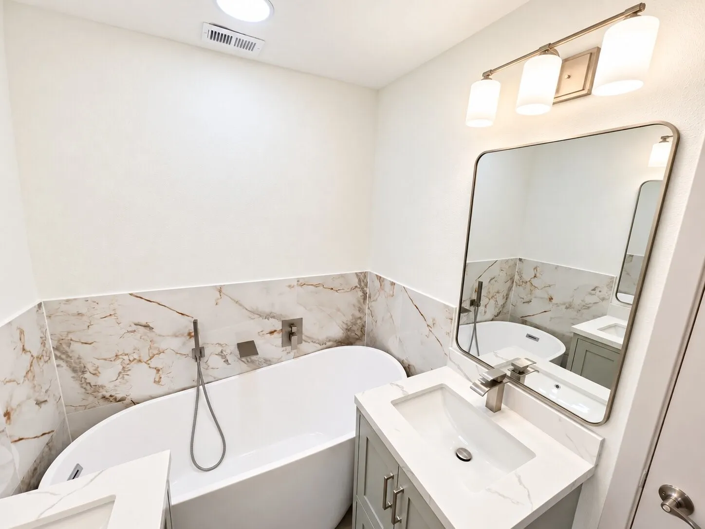 Freestanding soaking tub featuring wall-mounted chrome fixtures, light-toned stone-finish wall tile, vanity with stone countertop and rectangular undermount sink, and minimalist three-light vanity fixture