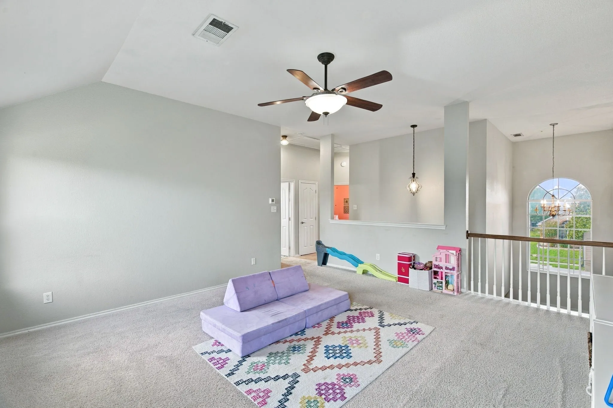 Vaulted ceilings, neutral carpeting, a central ceiling fan with integrated lighting, a half-wall partition with pendant lighting, and an arched window with a wooden railing