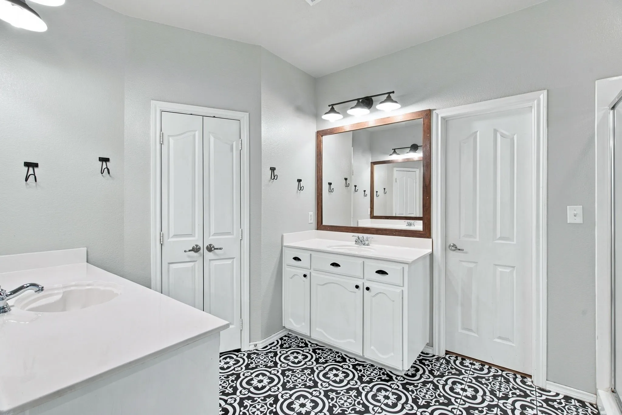 Bathroom featuring dual white vanities with solid-surface countertops, pattern-tiled flooring, three-light wall-mounted fixtures, and wood-framed mirrors