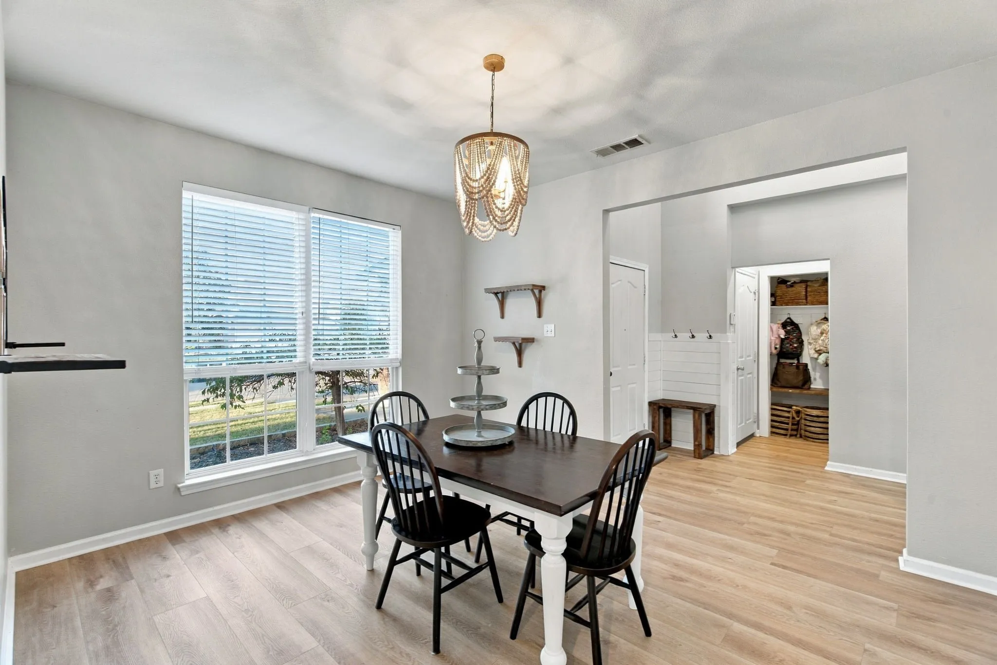 Wood-finish flooring, beaded chandelier, large windows with horizontal blinds, built-in wall shelving, and shiplap-style wall accent with coat hooks