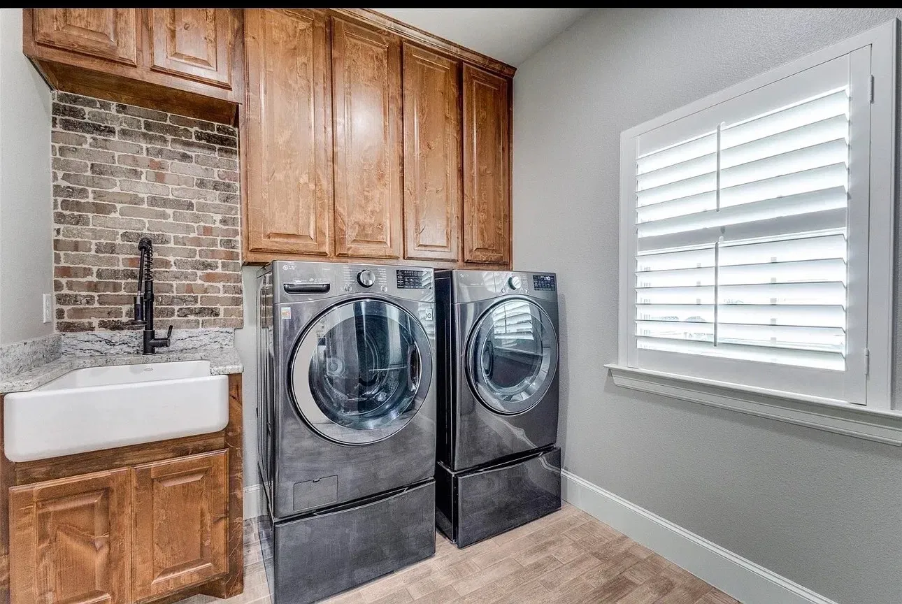 Built-in stained wood cabinetry, farmhouse-style apron sink, brick-tiled backsplash, stone countertop, and white plantation shutters