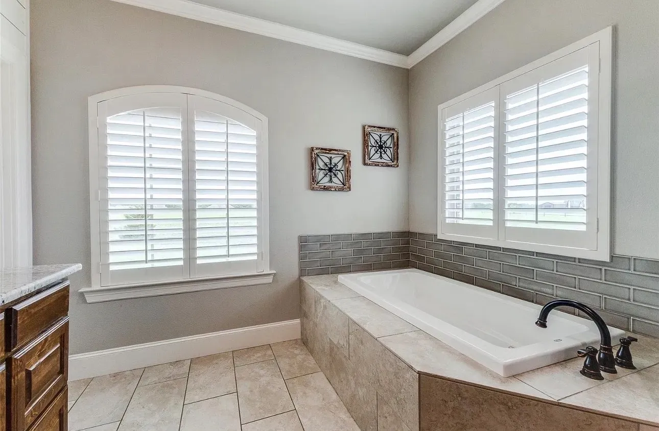 Built-in soaking tub with a tiled surround, arched window featuring white plantation shutters, and subway tile backsplash
