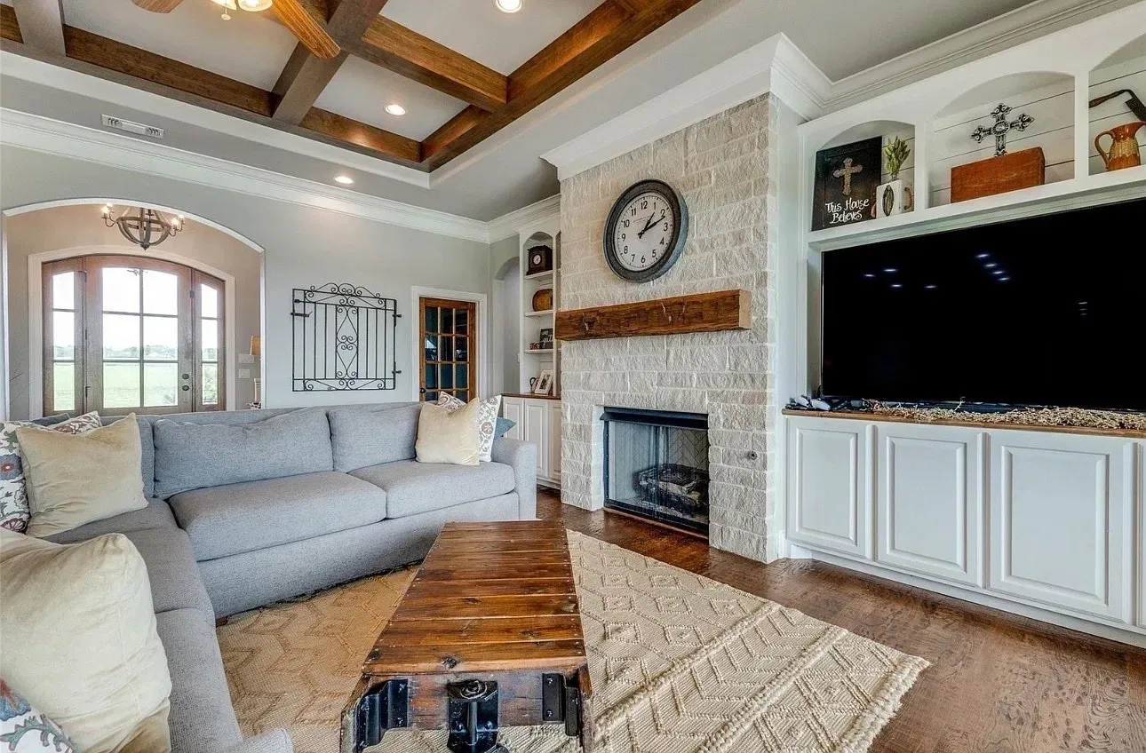 Living area featuring a coffered ceiling with exposed beams, natural stone fireplace surround, heavy timber mantel, and custom built-in cabinetry with integrated shelving