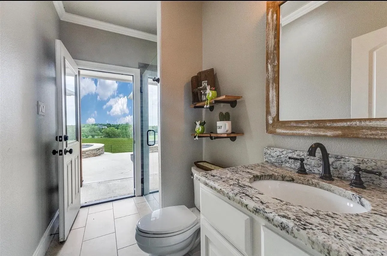 Stone-topped vanity with an oval undermount sink, oil-rubbed bronze faucet, wood-framed mirror, light-toned tile flooring, and direct access to an outdoor patio