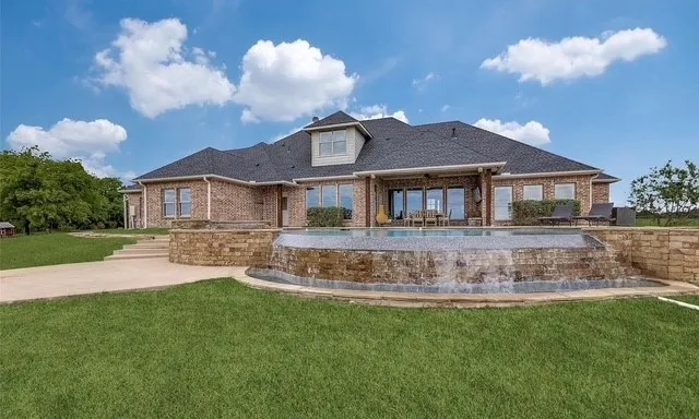 Brick exterior residence featuring a covered patio, an infinity-edge pool with a stone water feature, and expansive lawn grounds