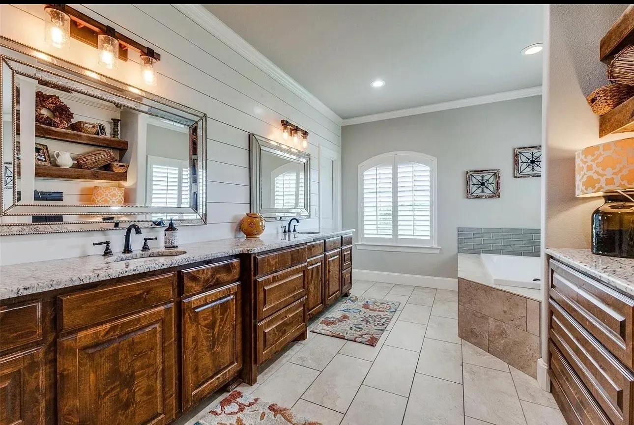 Dual-sink vanity with stone countertops, horizontal shiplap wall detailing, tile flooring, built-in soaking tub with stone surround, and an arched window featuring plantation shutters