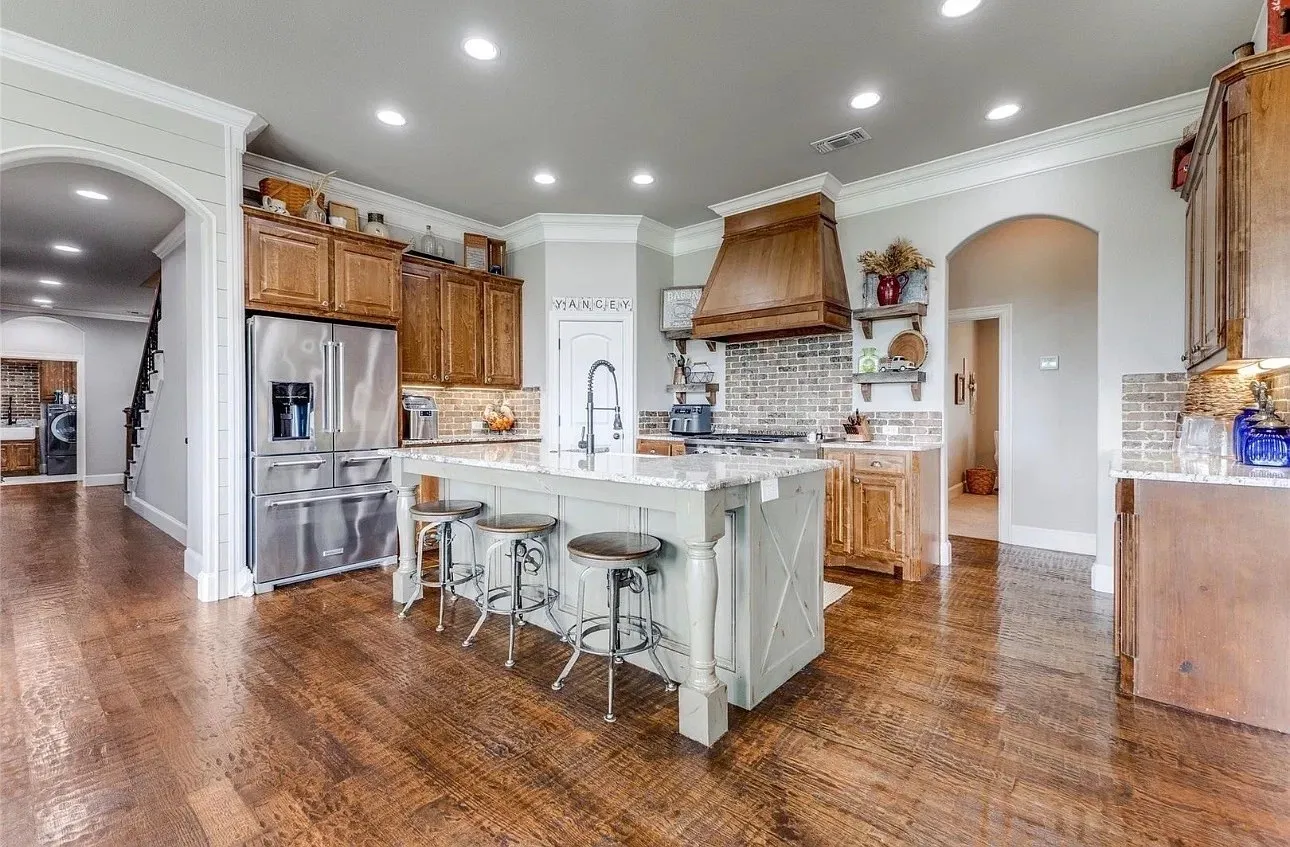 Kitchen featuring wood-finish flooring, a central island with stone-finish countertops, custom cabinetry, a wood range hood, and a brick backsplash