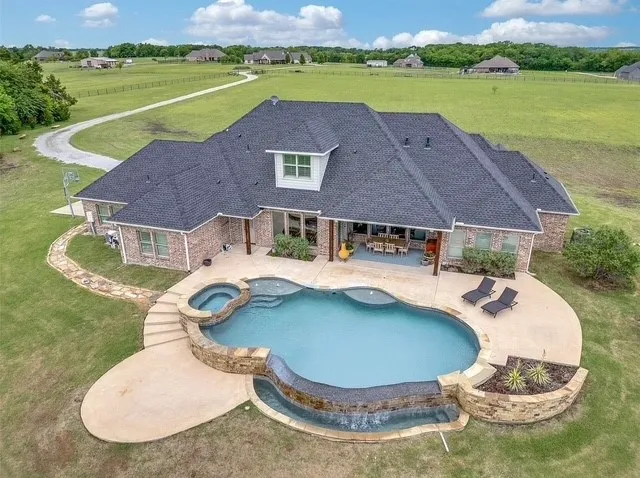 Brick exterior residence featuring a covered patio, a custom-shaped swimming pool with stone accents, a raised spa, and an expansive lawn
