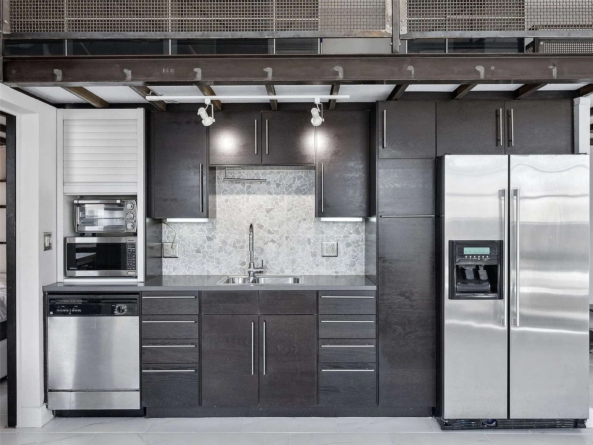 Kitchen featuring dark wood cabinetry, stainless steel appliances, mosaic tile backsplash, solid-surface countertops, and industrial-style exposed steel ceiling beams