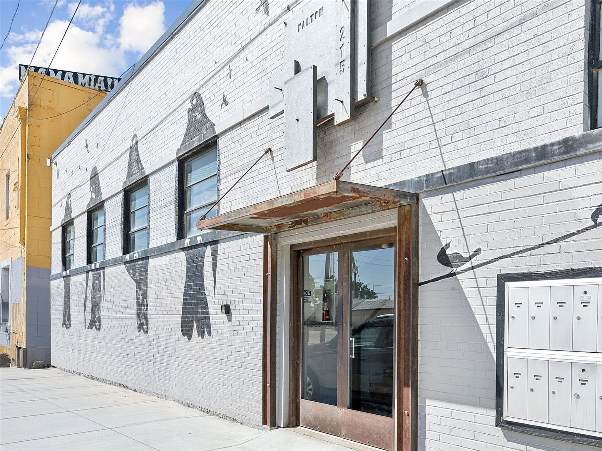 Painted brick exterior featuring industrial-style metal awning, glass and metal entrance doors, multi-unit wall-mounted mailbox system, and high-contrast mural detail