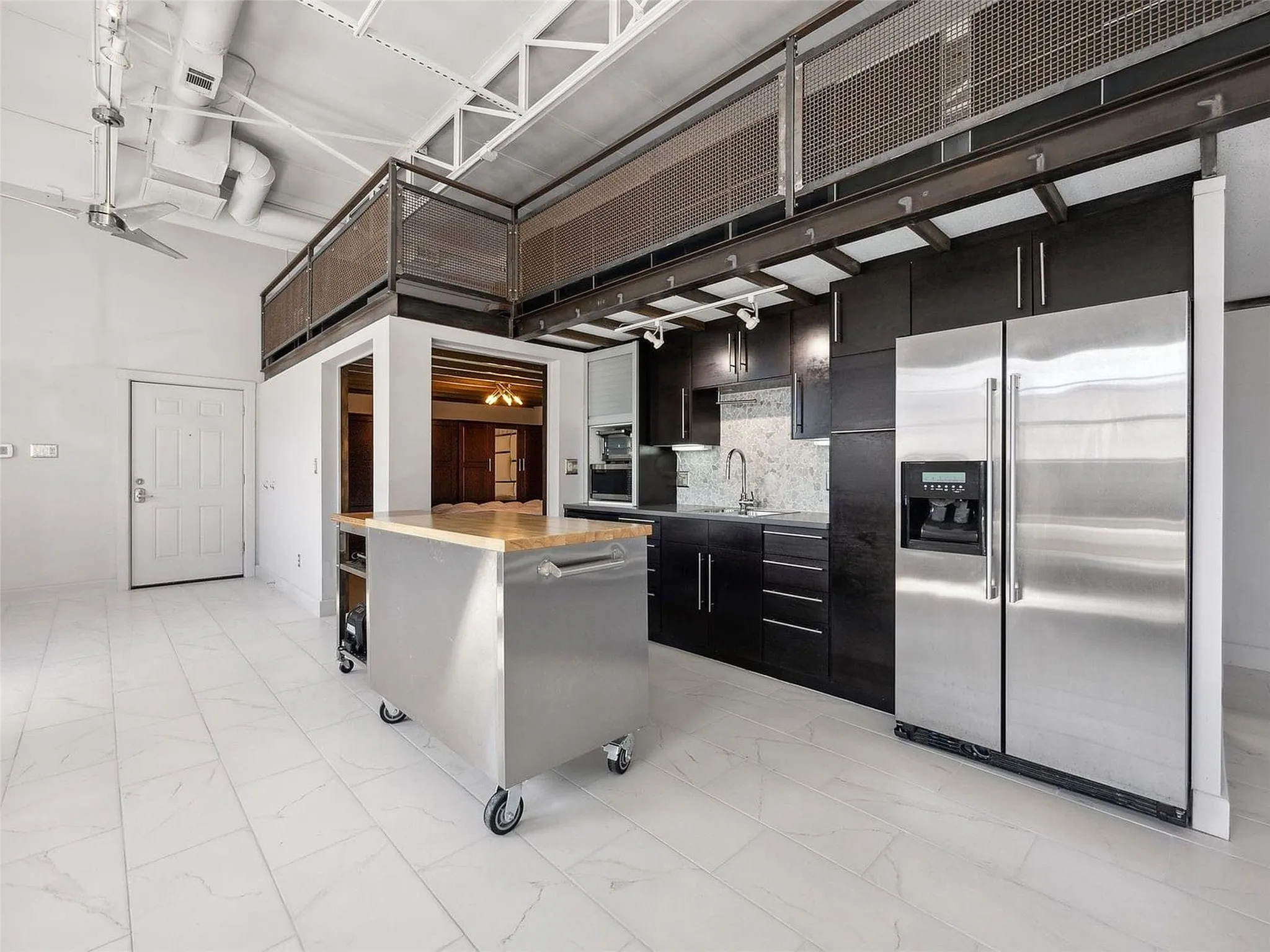 Industrial-style loft featuring high ceilings with exposed trusses, dark wood cabinetry, a stainless steel kitchen island, tiled flooring, and a metal-mesh mezzanine railing