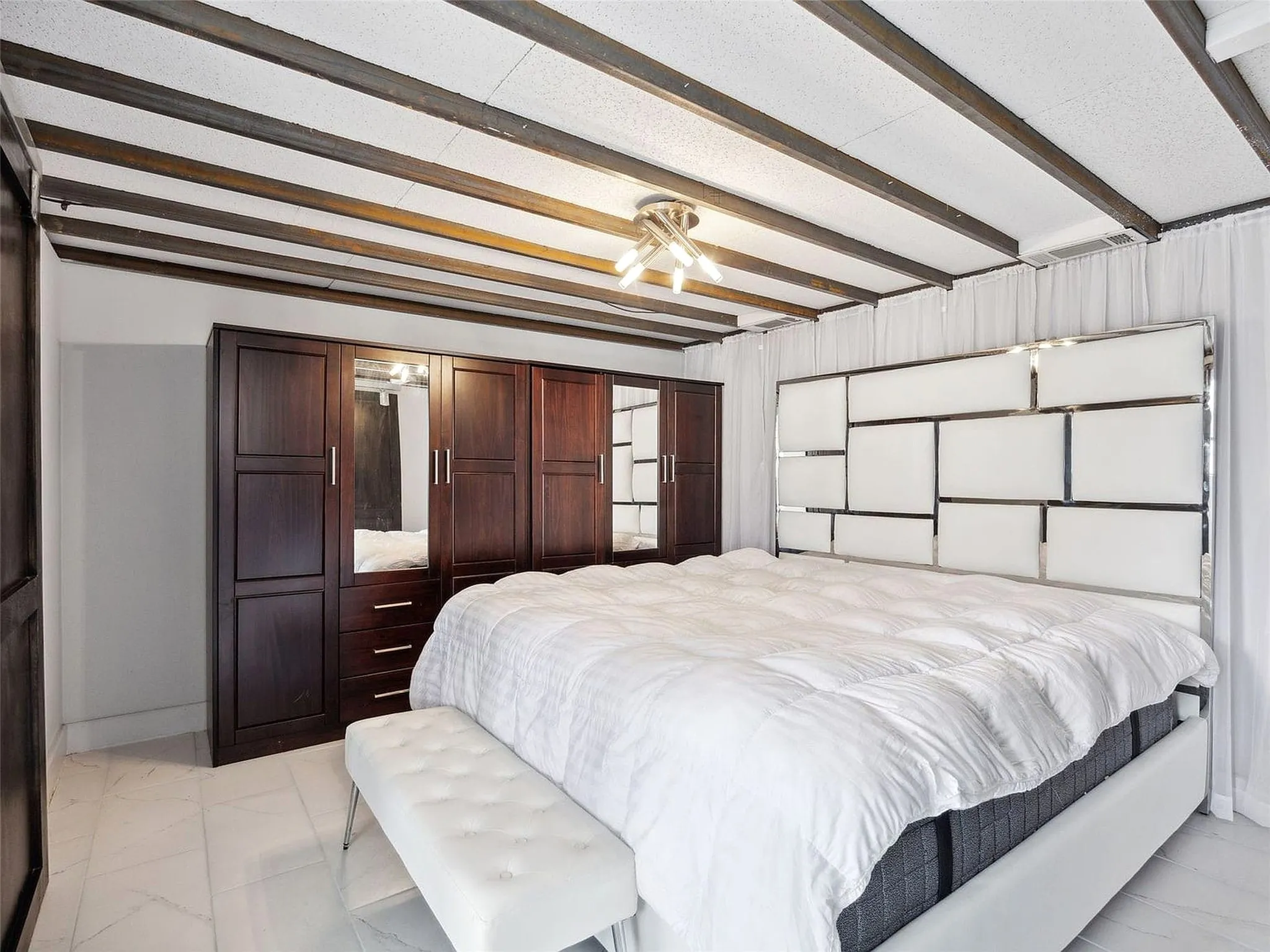 Primary bedroom suite featuring exposed beam ceiling with recessed lighting, dark wood built-in cabinetry and wardrobe system, and a geometric accent wall with white paneling