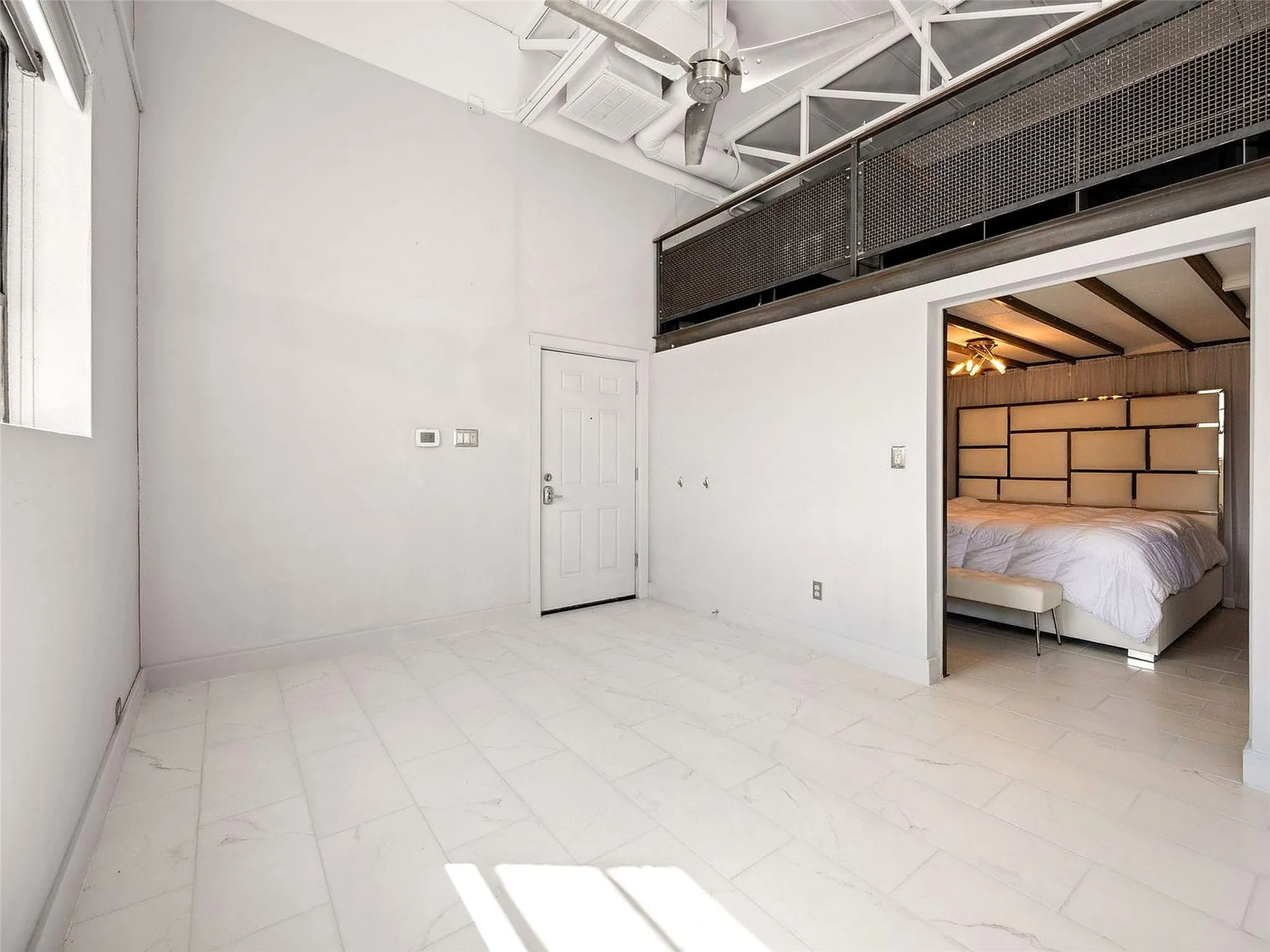 High-ceiling industrial interior featuring exposed rafters, a metal mesh loft railing, ceiling-mounted paddle fan, and large-format tile flooring