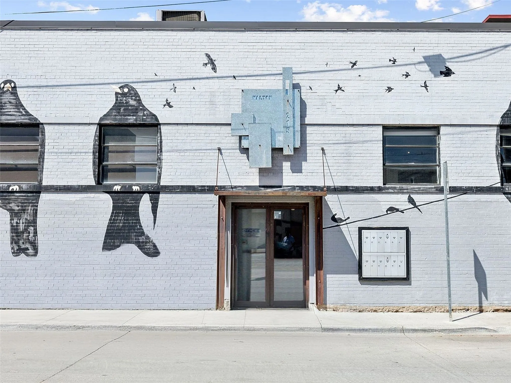 Painted masonry exterior featuring mural artwork, metal-frame glass entry door, multi-pane industrial-style windows, and mounted metal mail distribution station