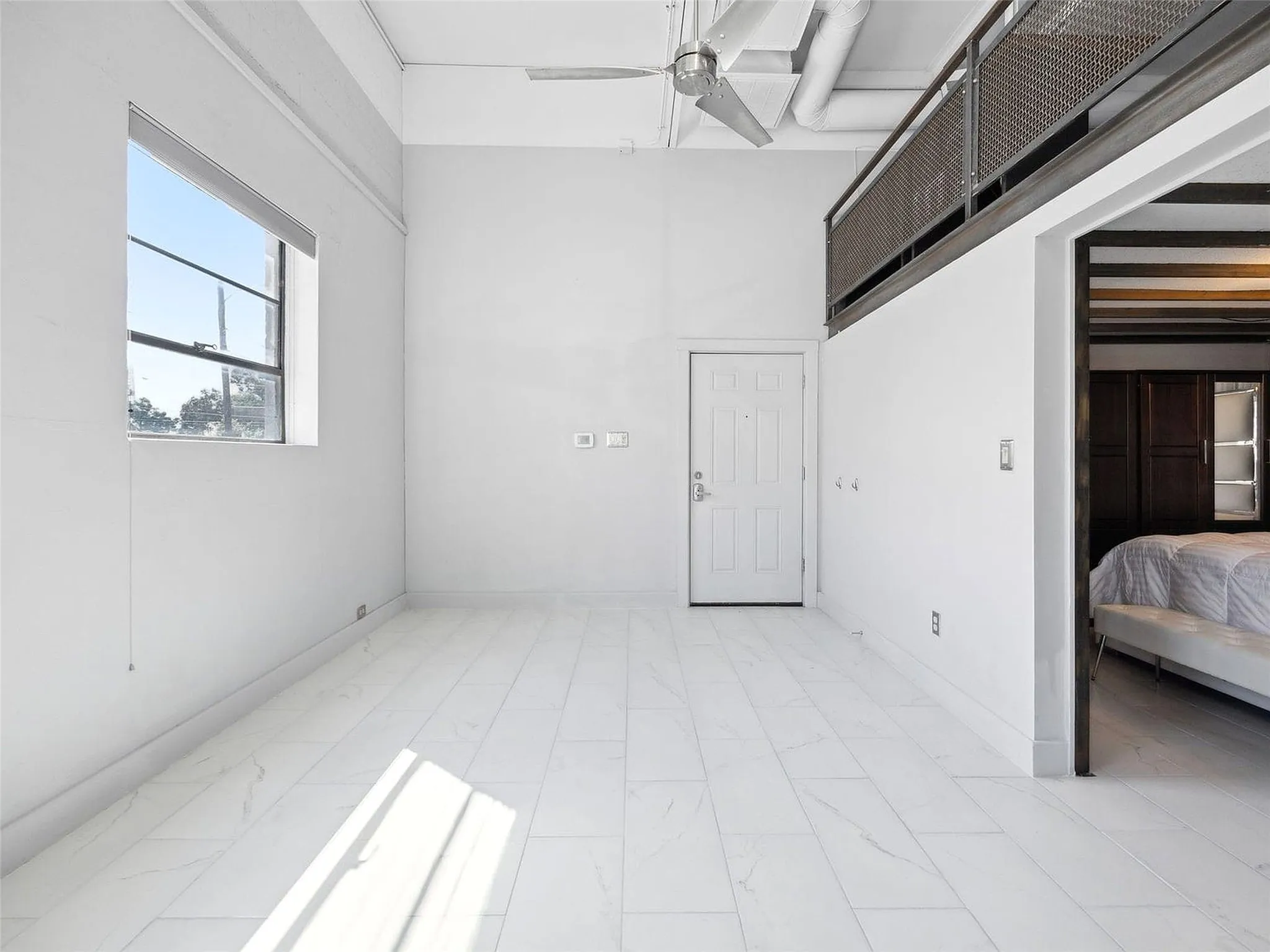 High-ceiling industrial loft featuring white tile flooring, exposed ductwork, a metal mesh mezzanine railing, and a ceiling fan