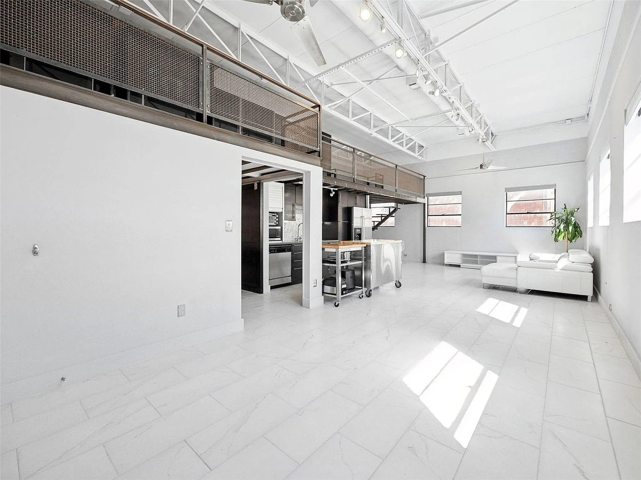 Industrial-style interior featuring high vaulted ceilings with exposed white trusses, large-format tile flooring, a mezzanine level with decorative metal mesh railing, and track lighting