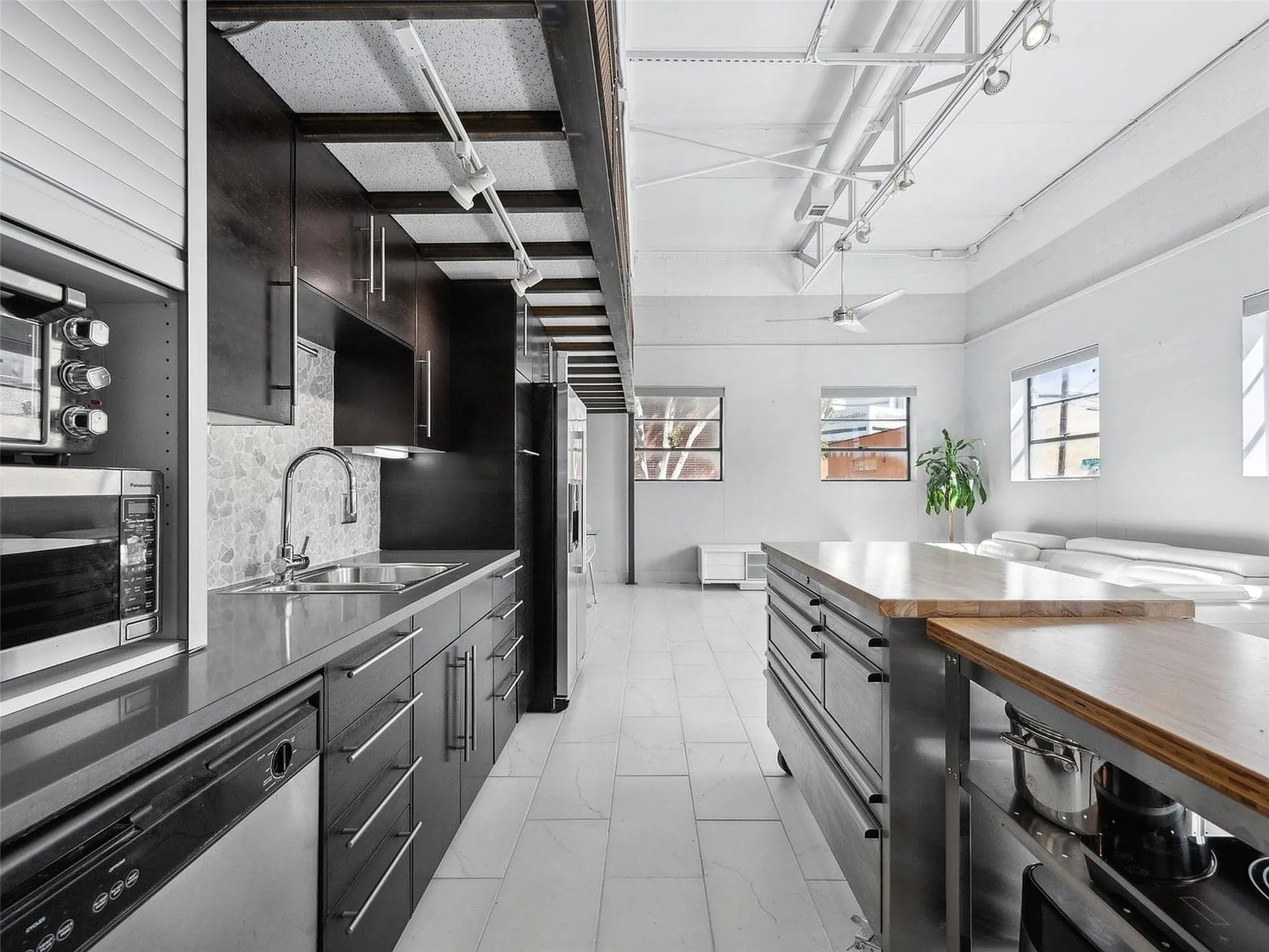 Industrial-style kitchen featuring sleek dark cabinetry, quartz countertops, tile flooring, exposed structural beams with track lighting, and a dual-level kitchen island with a wood-finish surface