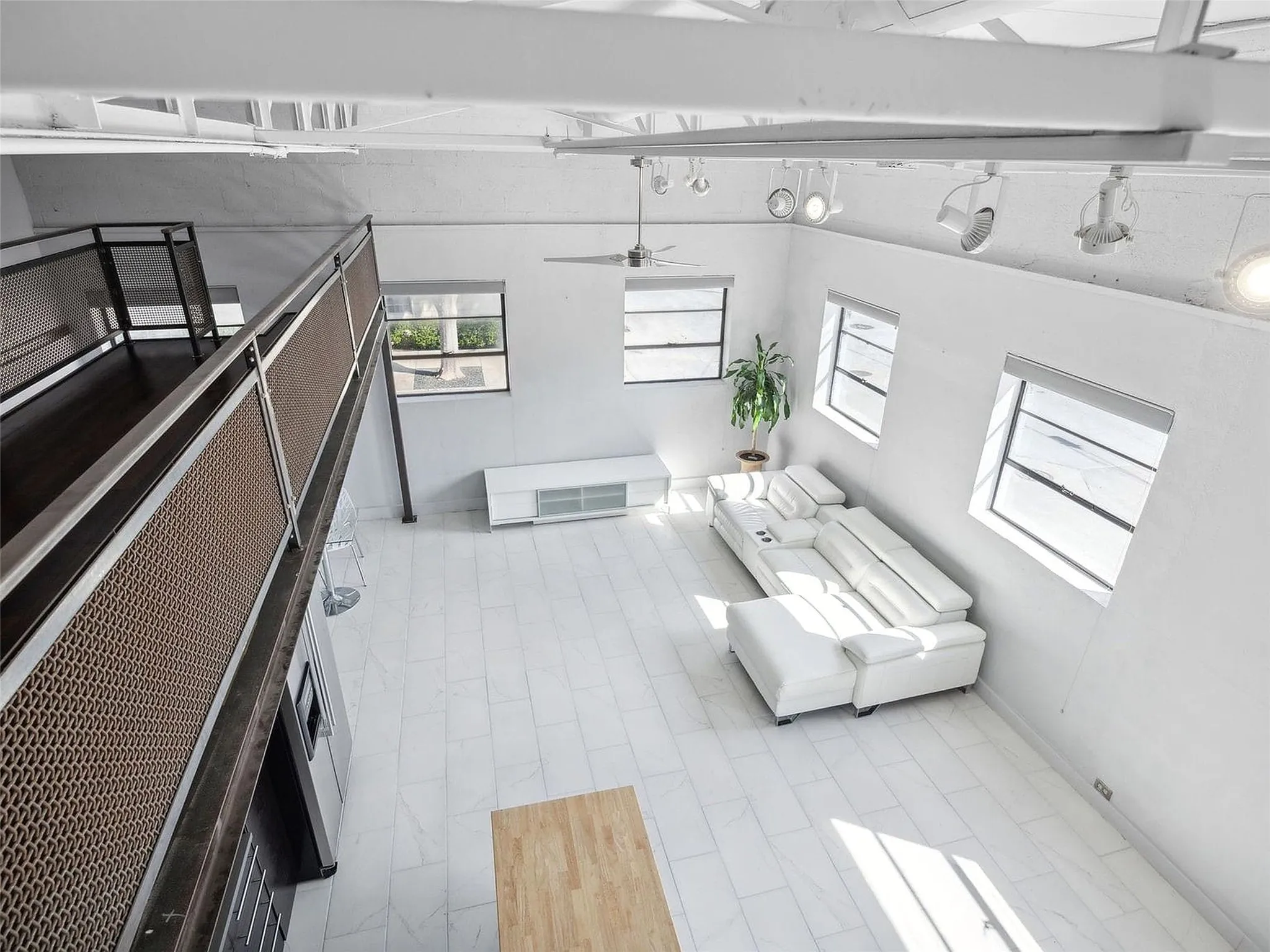Industrial-style loft interior featuring exposed white-painted truss ceilings, large multi-pane windows, polished tile flooring, and a metal mesh mezzanine railing