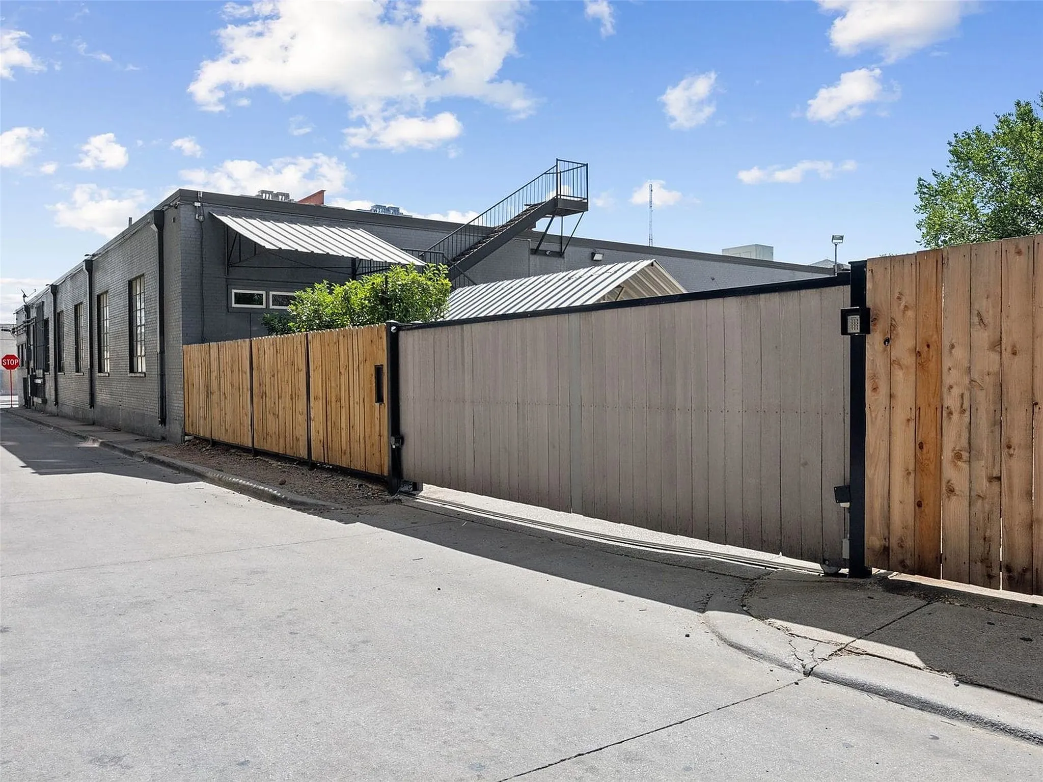 Exterior view featuring a motorized sliding wood-panel fence, dark-painted masonry siding, metal awning structures, and an exterior steel staircase