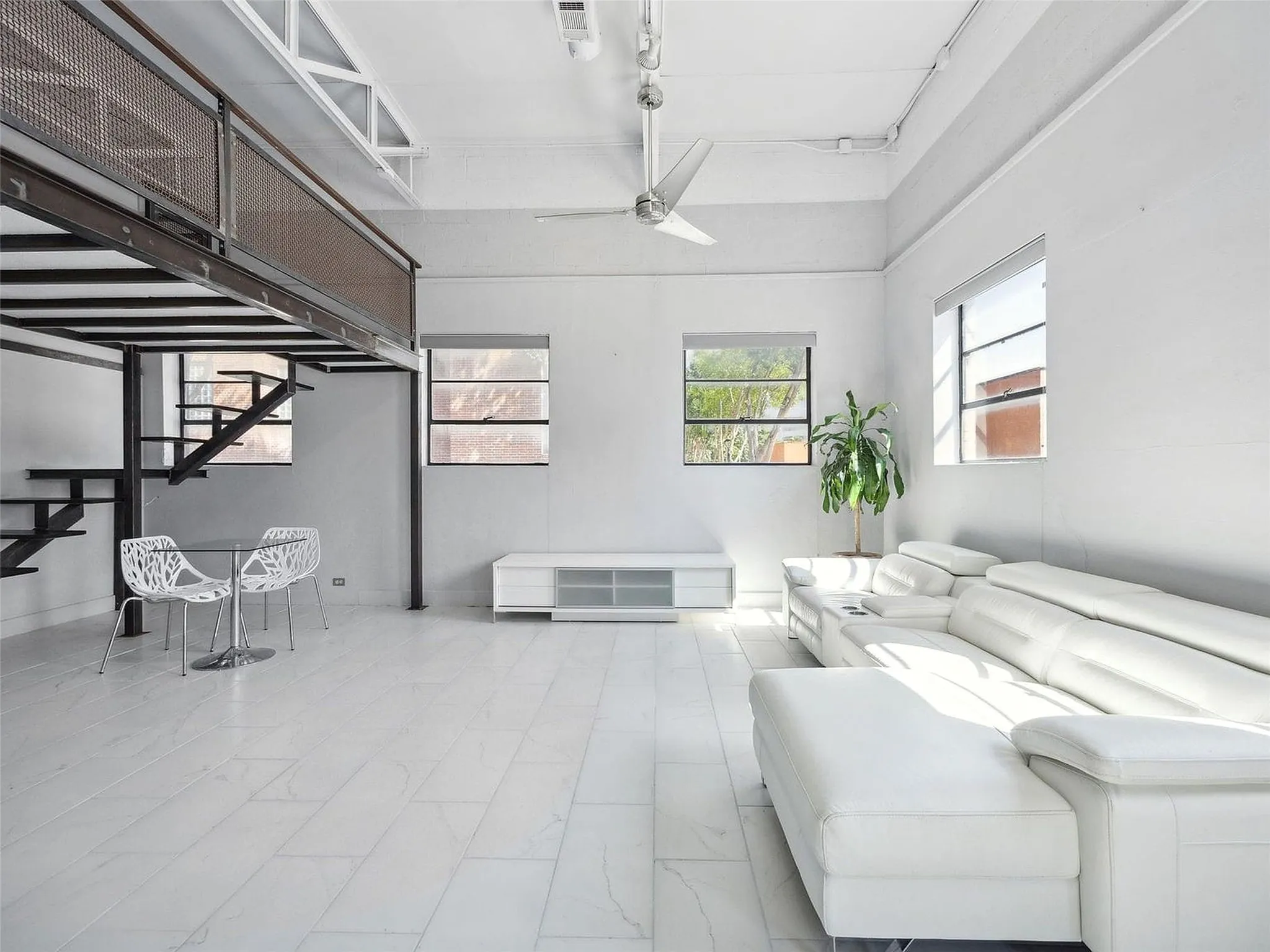 Industrial-style interior featuring high ceilings with exposed structural elements, a metal mezzanine with mesh railing, large windows, and neutral tile flooring