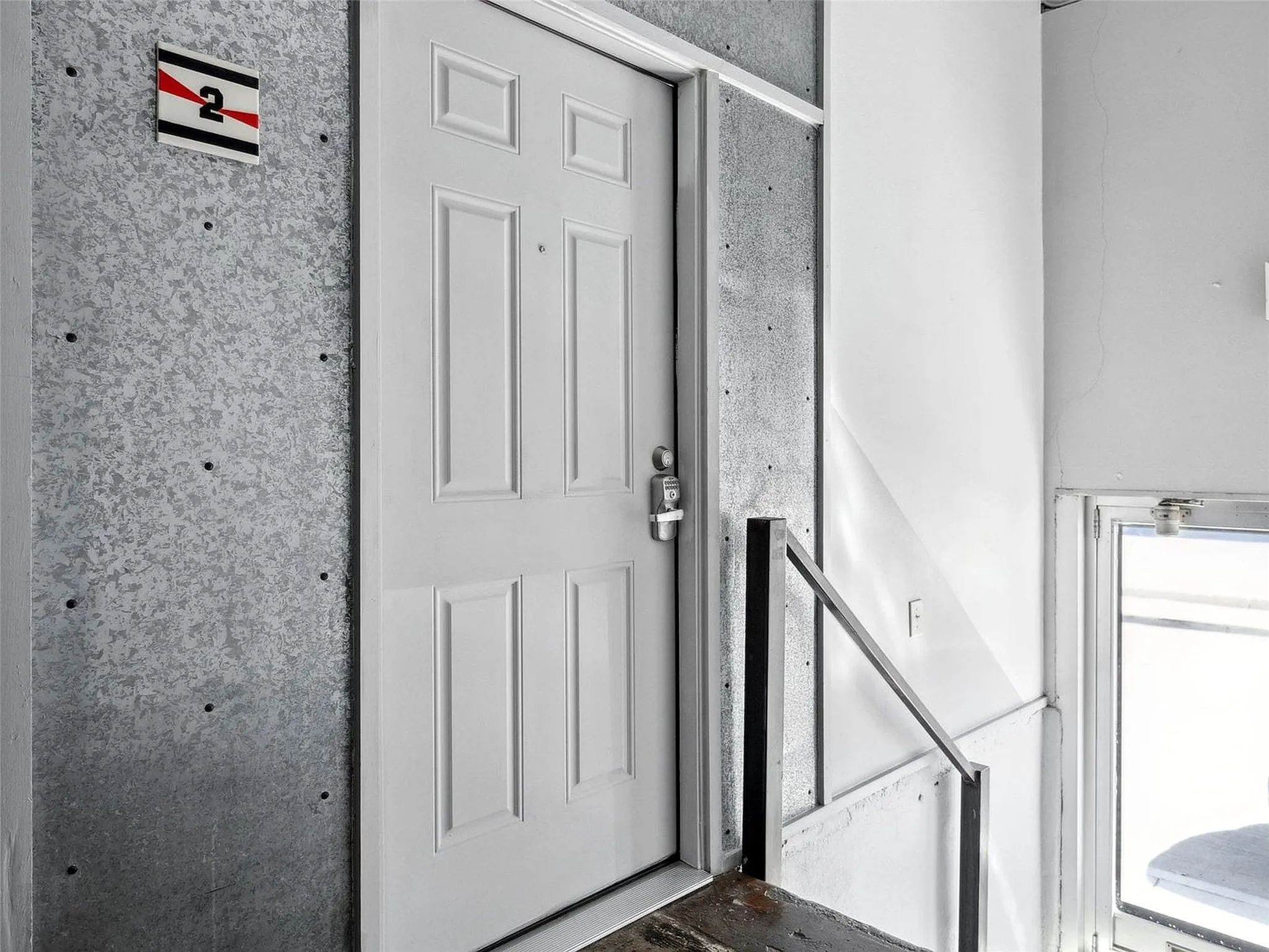 Four-panel entry door featuring a brushed metal deadbolt and integrated lock system, set against exposed concrete wall panels with visible mounting hardware and a metal handrail
