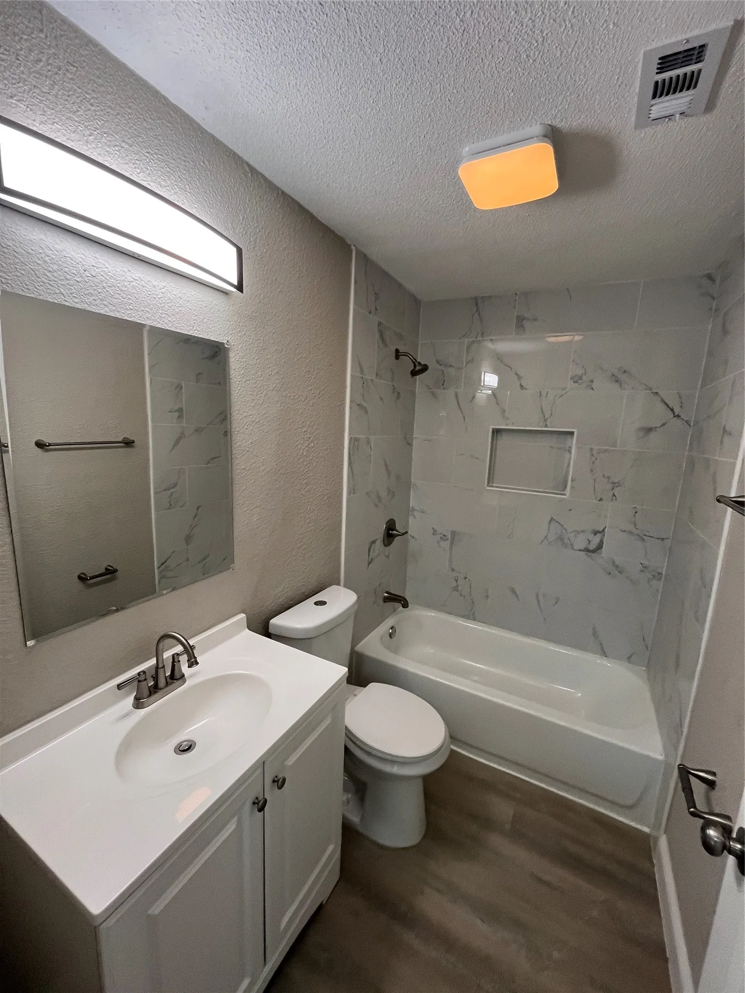 Bathroom featuring a vanity with a white countertop and oval under-mount sink, wood-finish flooring, and a tub-shower combination enclosed by large-format, light-toned tile with integrated shelving