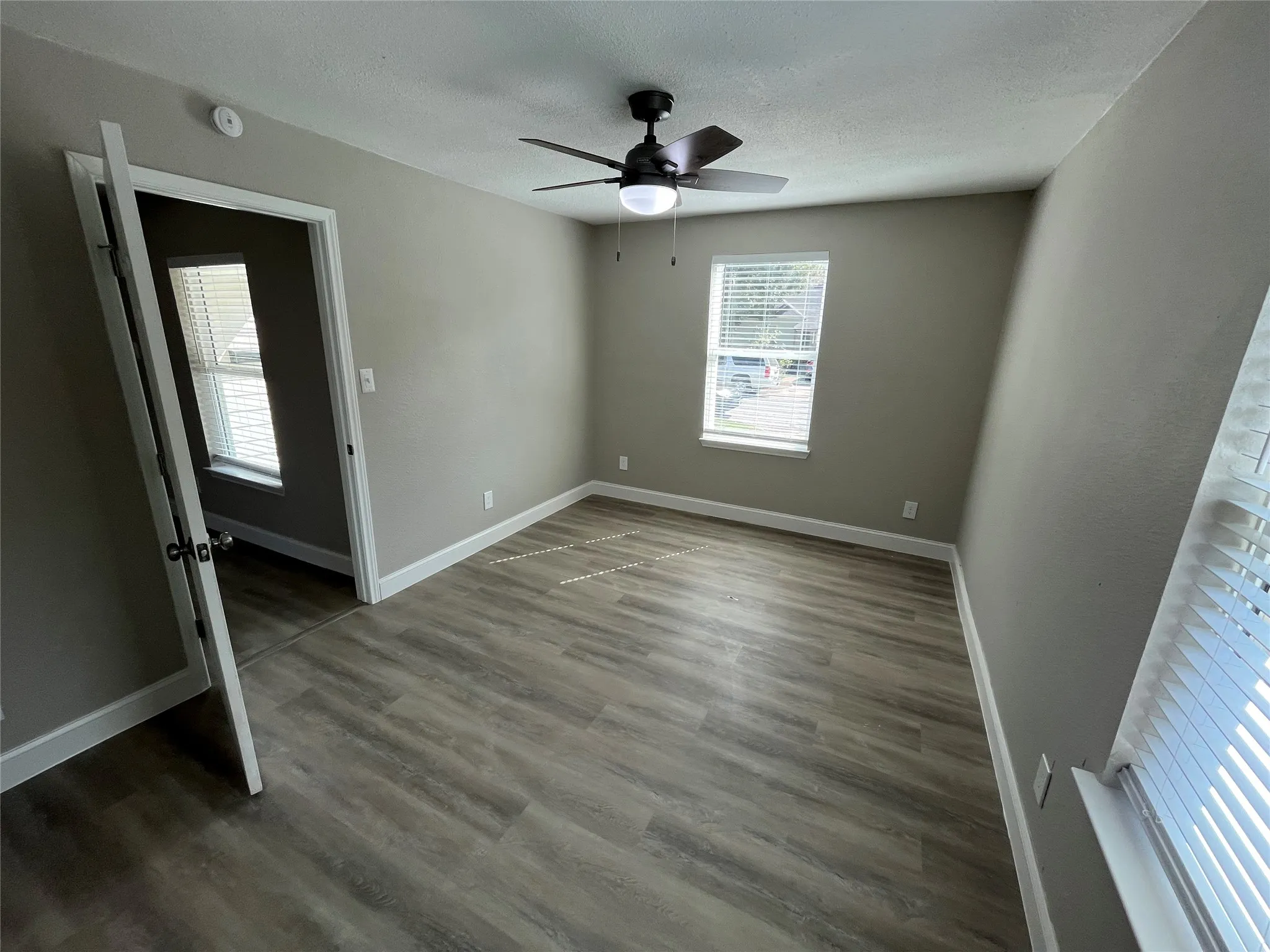 Neutral-toned interior featuring wood-finish flooring, dual windows with fitted blinds, and a central ceiling fan with integrated lighting