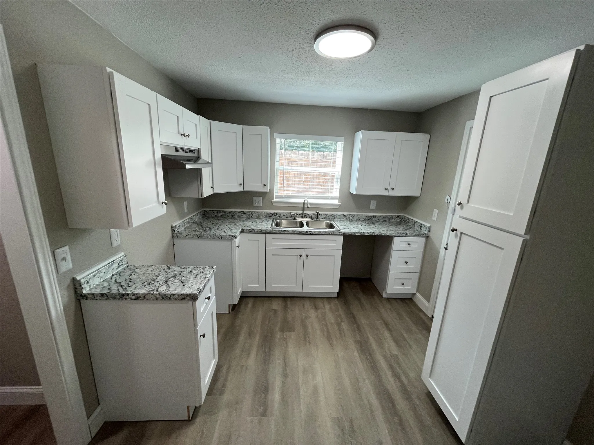Kitchen featuring white shaker-style cabinetry, patterned stone-finish countertops, stainless steel dual-basin sink, and wood-finish flooring