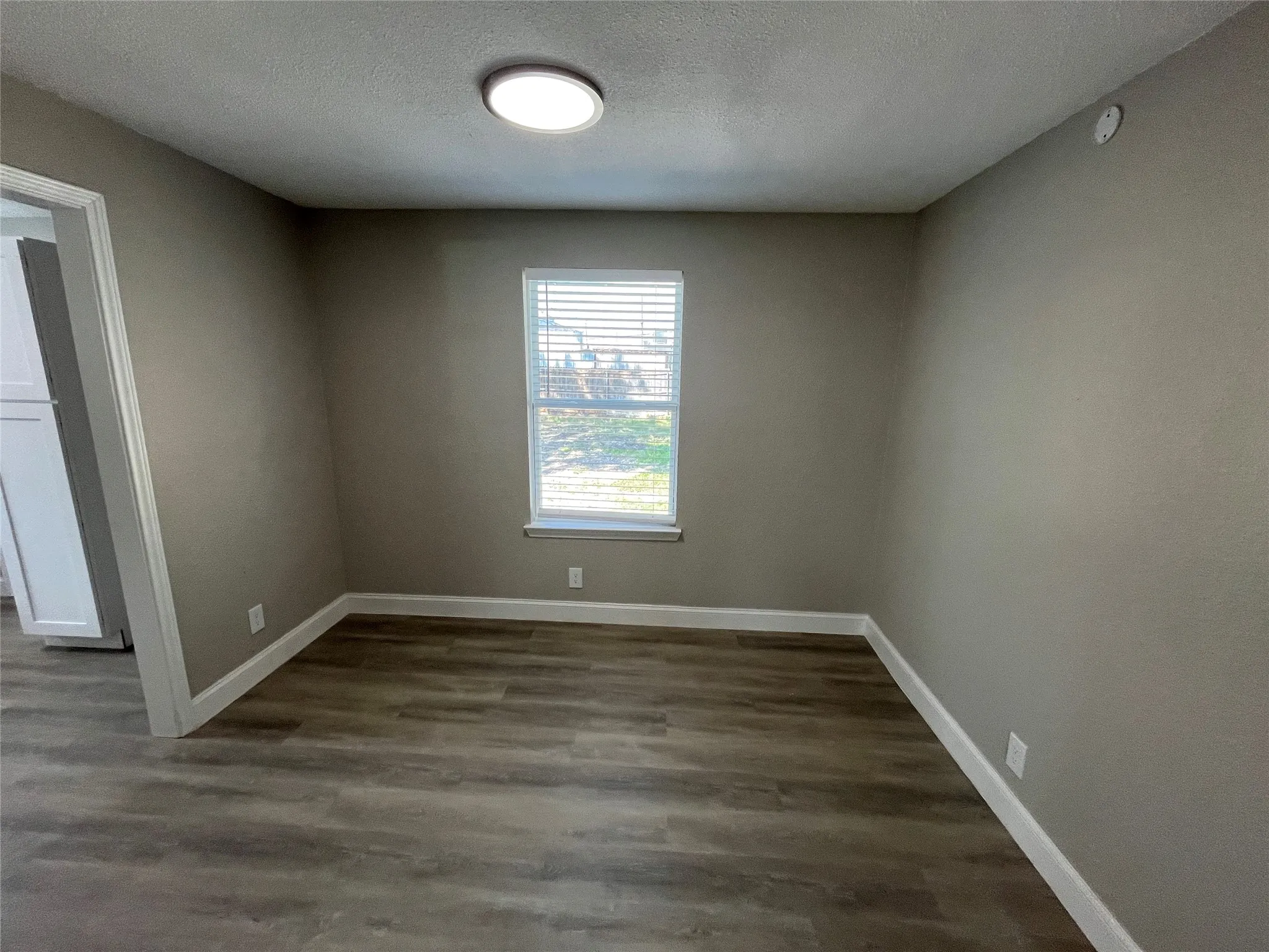 Neutral-toned interior room featuring wood-finish flooring, a single window with integrated horizontal blinds, and a circular flush-mount ceiling light fixture