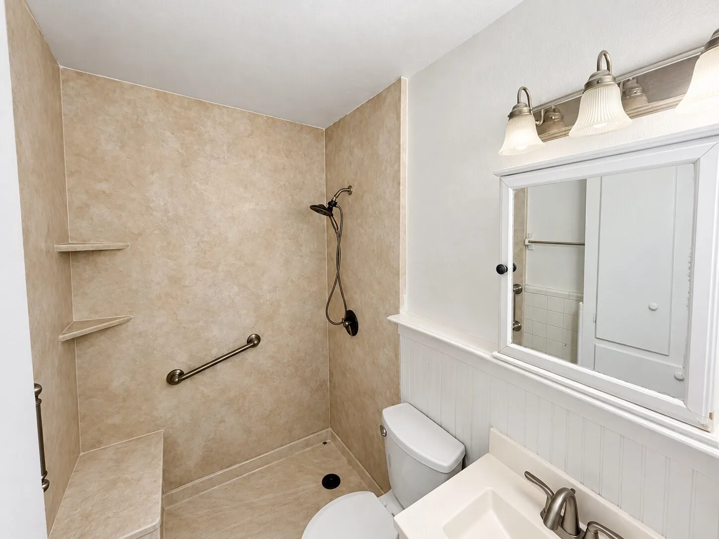 Walk-in shower with integrated bench seating, solid-surface surround, and dual shower heads, complemented by a vanity with an integrated basin, beadboard wainscoting, and multi-bulb wall lighting