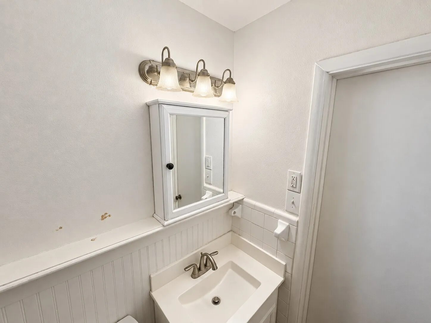 Wall-mounted medicine cabinet, integrated basin vanity, beadboard wainscoting, and three-light brushed-finish vanity fixture