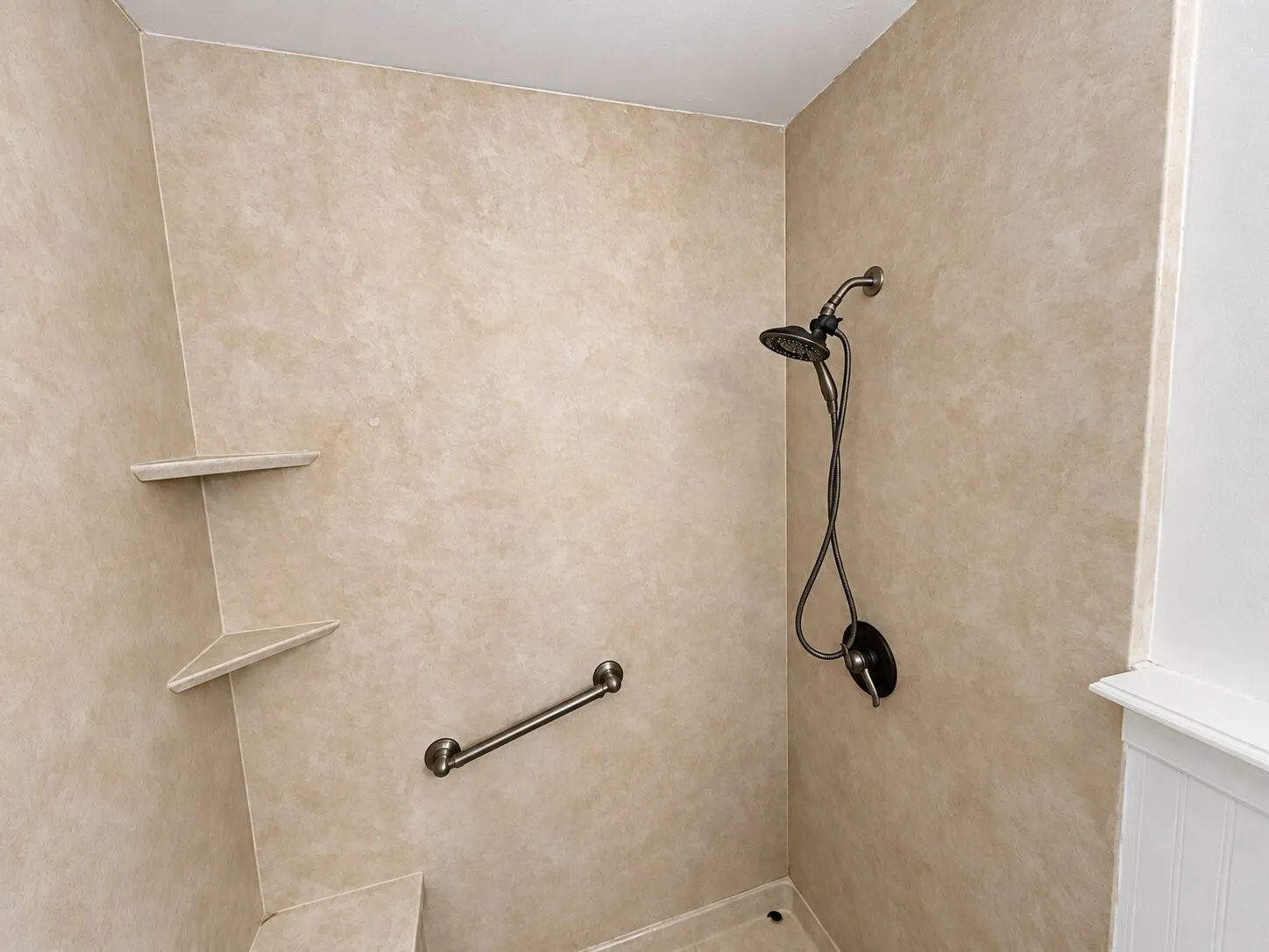 Shower stall featuring beige solid-surface surround panels, integrated corner shelving, a handheld shower fixture, and a horizontal metal grab bar