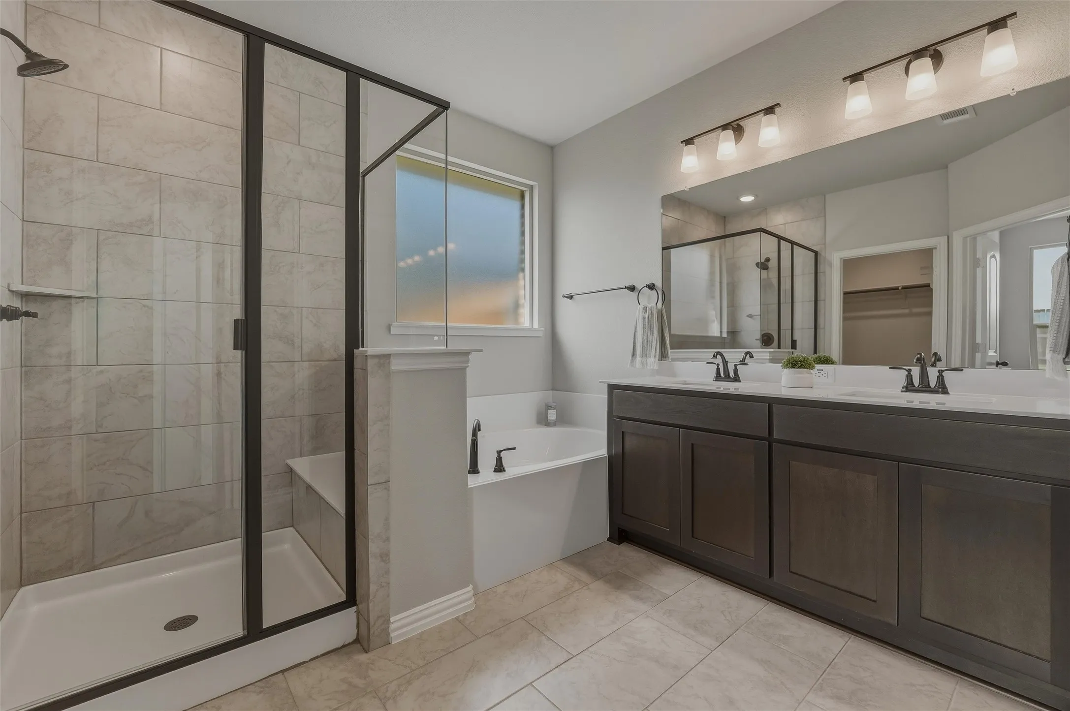 Spacious bathroom featuring a dual vanity with dark wood-finish cabinetry and undermount sinks, a large mirror, a separate soaking tub with black fixtures, and a glass-enclosed shower with tiled walls and a built-in bench