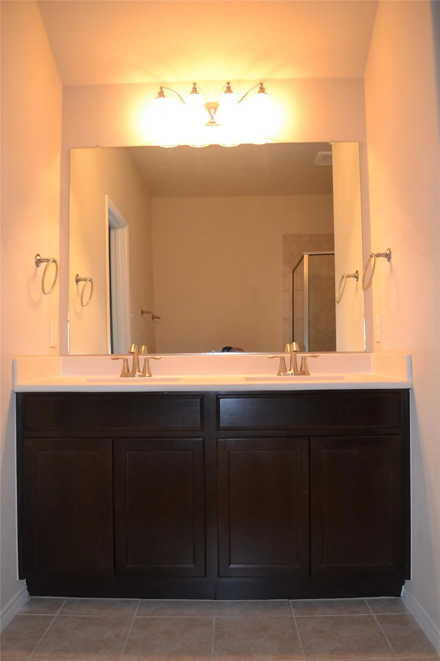 Dual-sink vanity featuring dark wood-finish cabinetry, solid surface countertop, metallic finish faucets, and a large-format vanity mirror with integrated light fixture