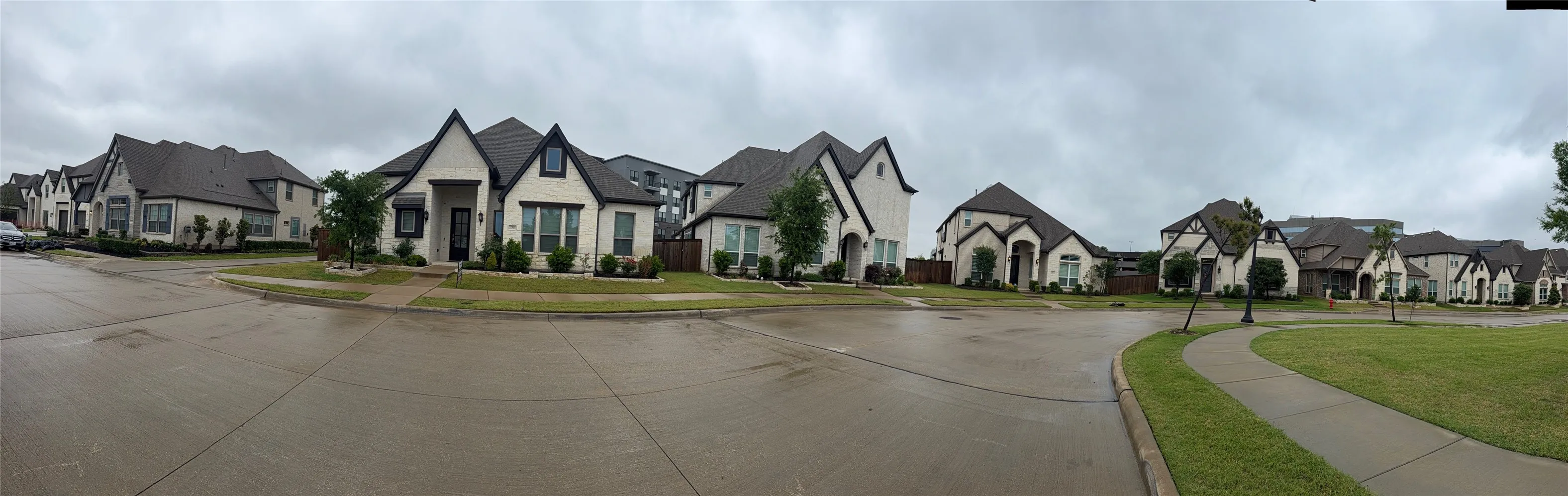 Street-level panoramic view of residential exteriors featuring multi-gabled rooflines, light-toned masonry facades, dark-trimmed window frames, and concrete driveways