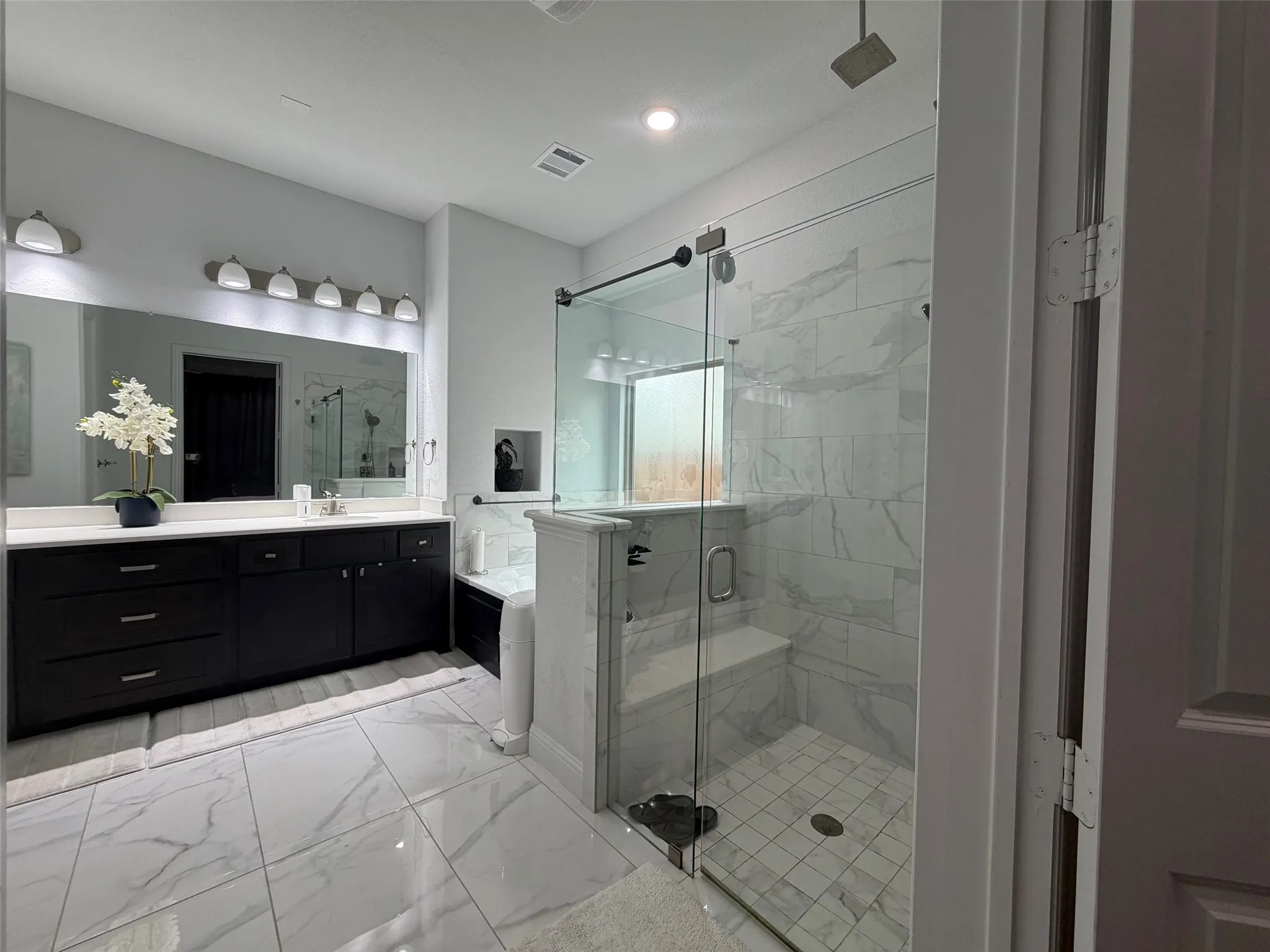 Bathroom featuring dark cabinetry with a light countertop, large-format tile flooring, a glass-enclosed shower with tiled walls and built-in bench, and a wall-to-wall mirror