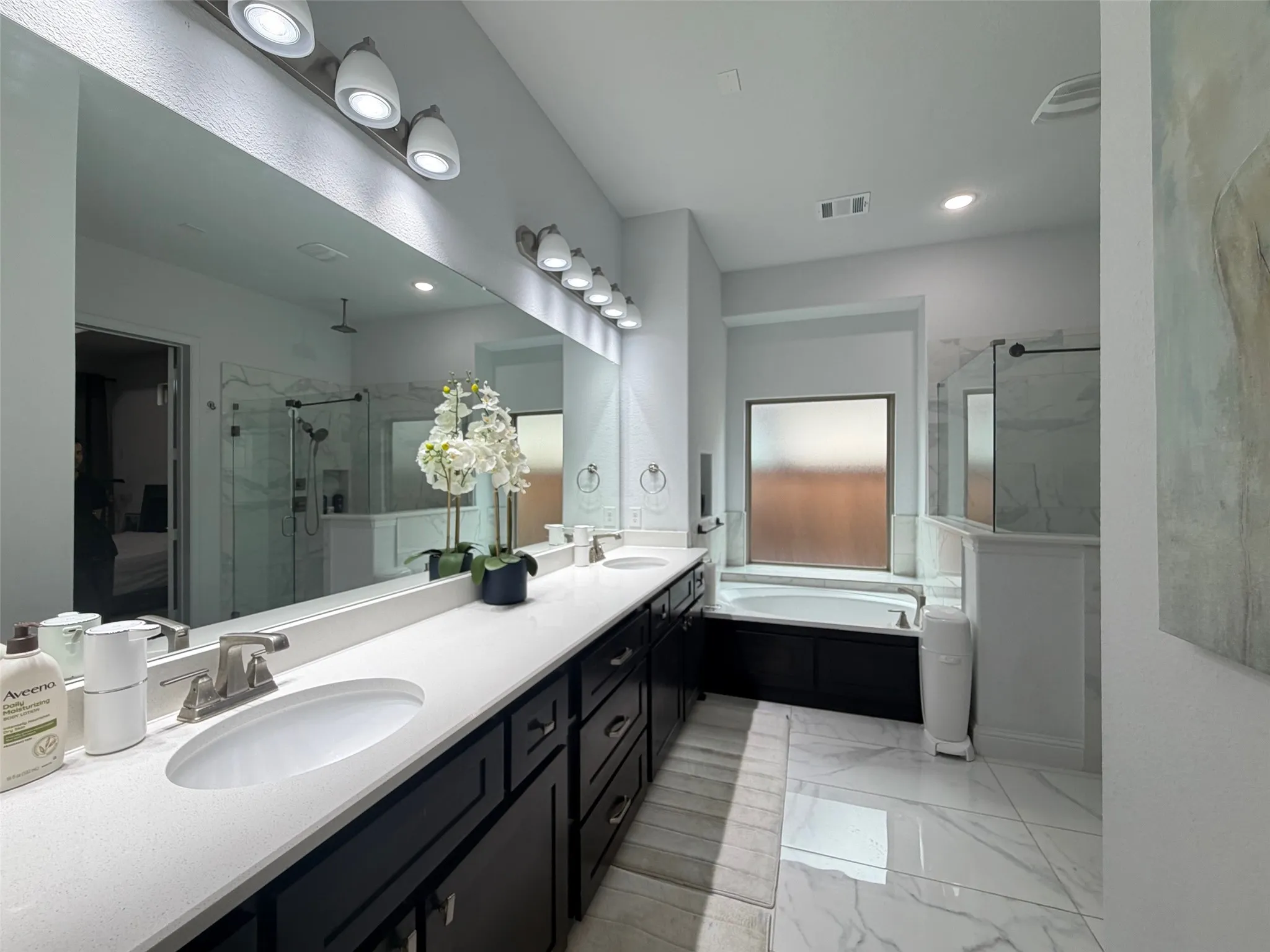 Dual-sink vanity with light-toned quartz countertops and dark cabinetry, large wall-mounted mirror, soaking tub, and walk-in glass-enclosed shower with tiled surround