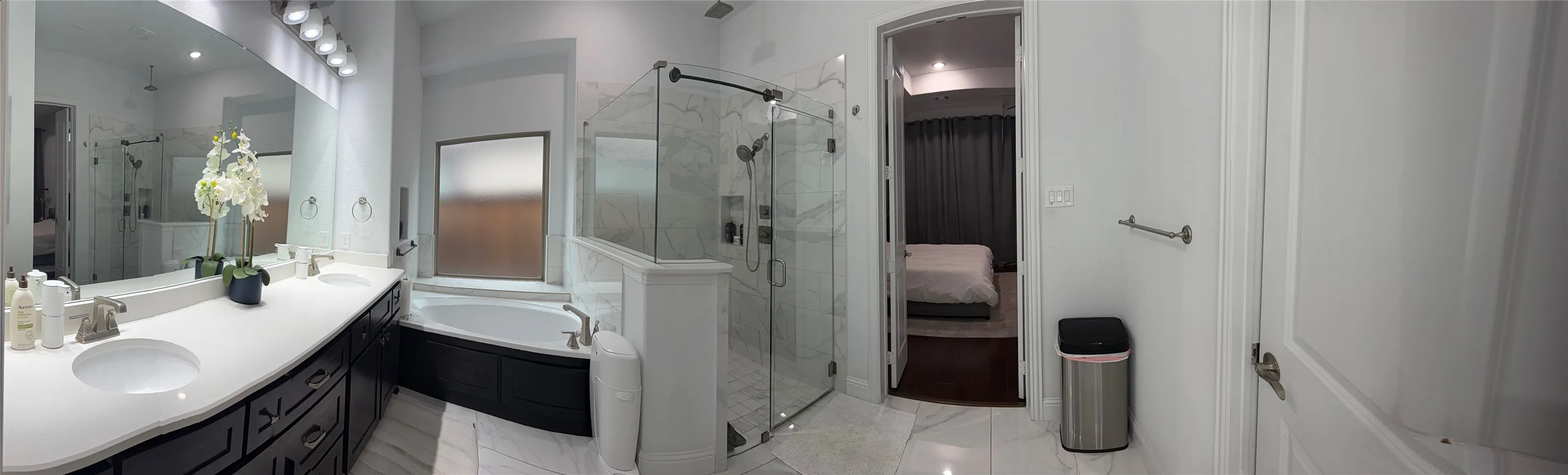 Double vanity with stone-style countertop and integrated sinks, deep soaking tub, glass-enclosed shower with built-in niche, and large wall-to-wall vanity mirror