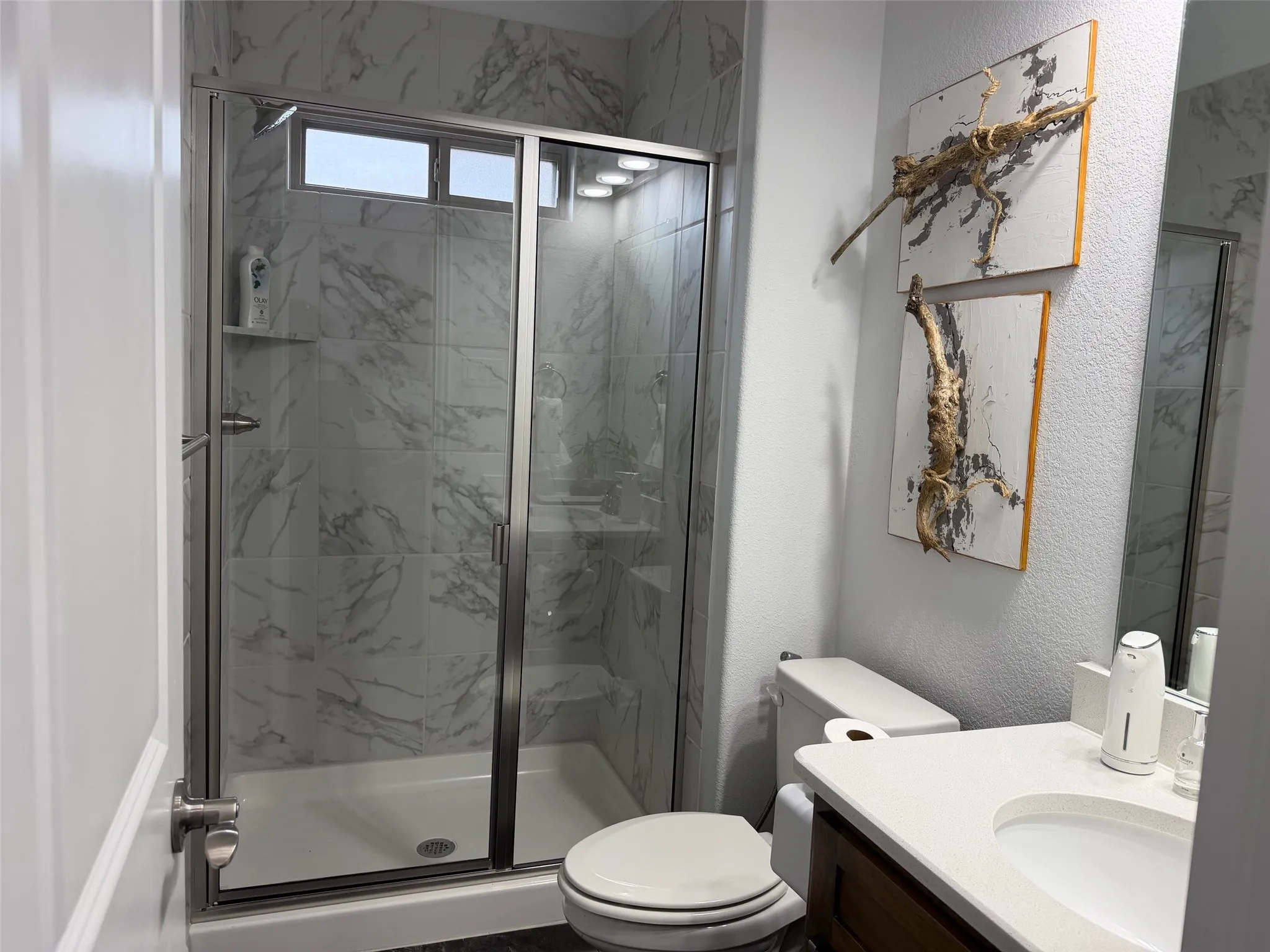 Walk-in shower with stone-finish tile surround and glass enclosure, vanity with integrated basin and light-toned countertop, and recessed ceiling lighting