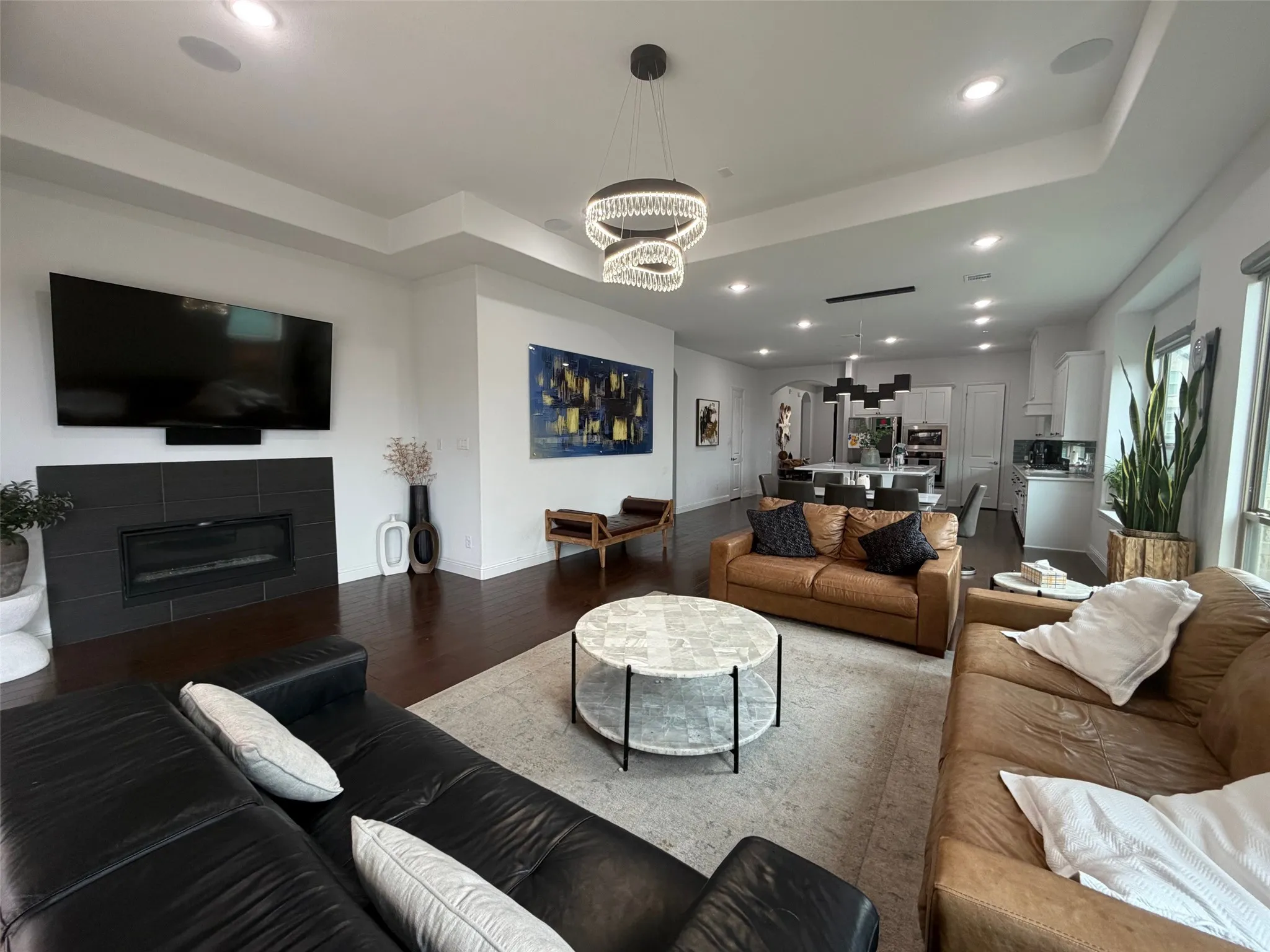 Open-concept living area featuring dark wood-finish flooring, a tray ceiling, a modern multi-tier crystal chandelier, recessed lighting, and a linear fireplace with a matte tile surround