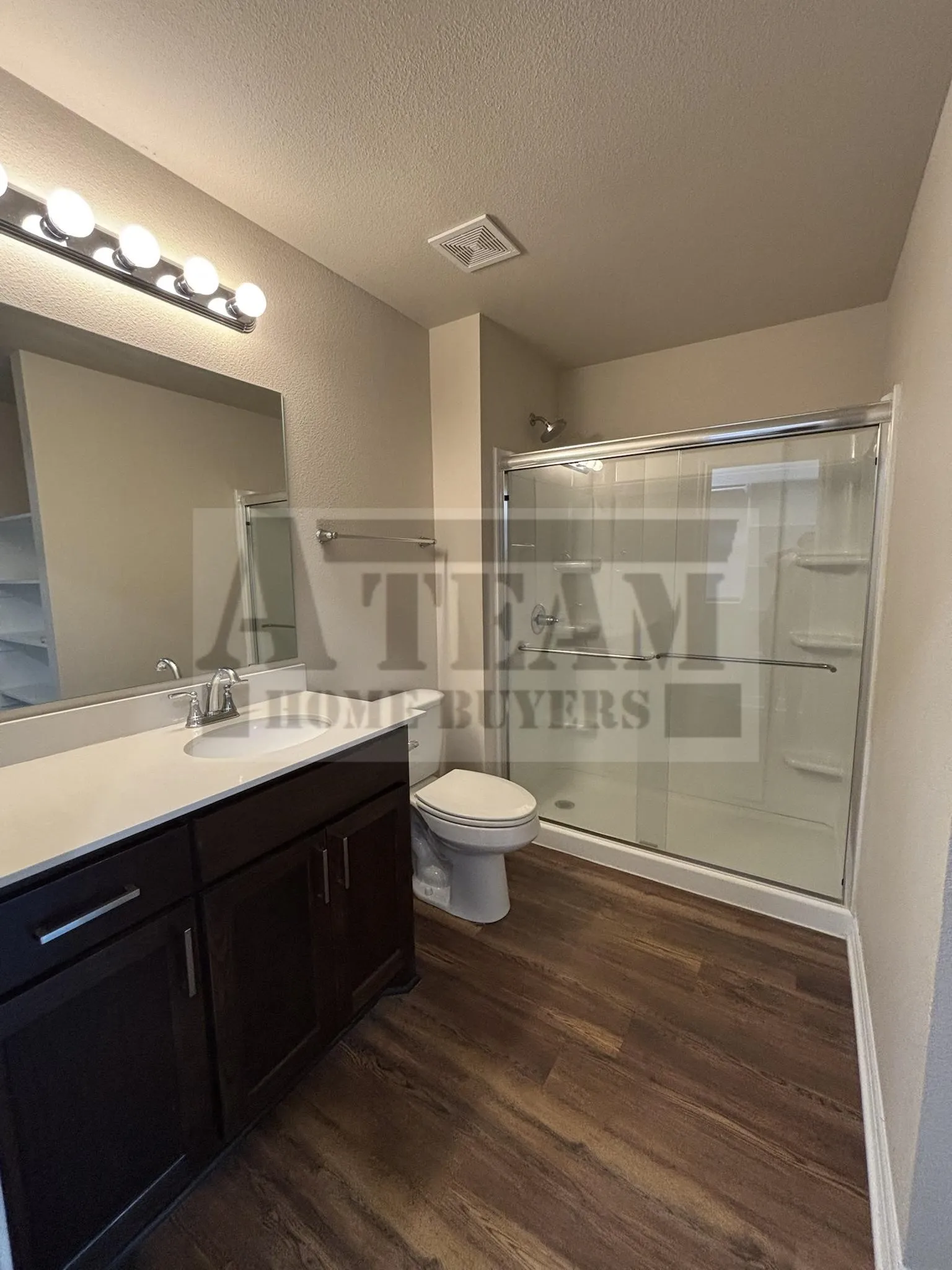 Dark wood-finish cabinetry, light-toned countertop with an integrated sink, polished hardware, wood-finish flooring, and a glass-enclosed shower stall