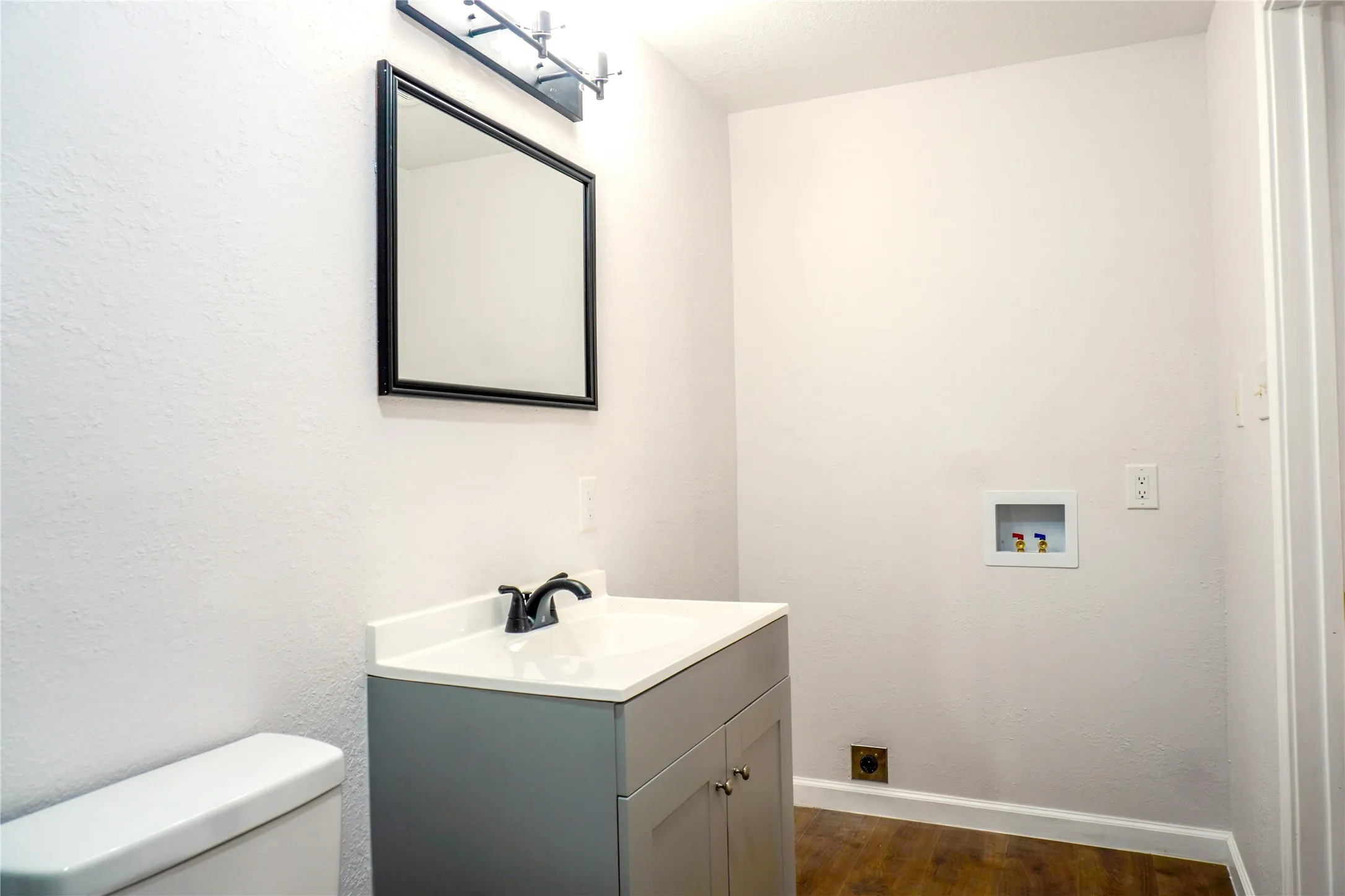 Vanity featuring a gray cabinet with white countertop, integrated sink, and black faucet, complemented by a framed rectangular mirror, overhead lighting, and wood-finish flooring