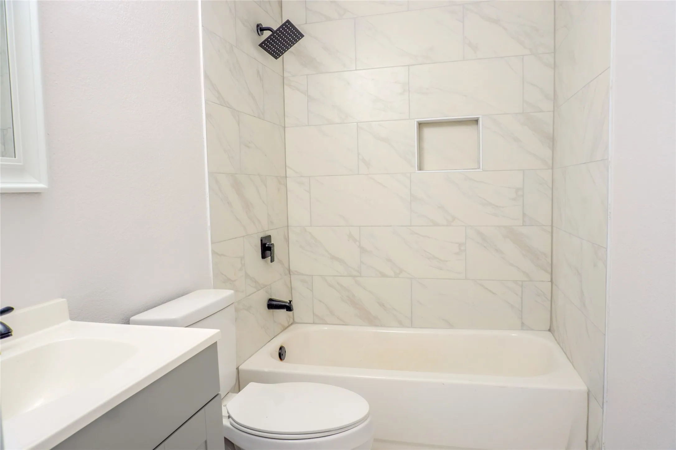 Bathroom featuring a shower-tub combination with light-toned wall tile, a built-in recessed niche, matte black fixtures, and a gray vanity with a white integrated sink