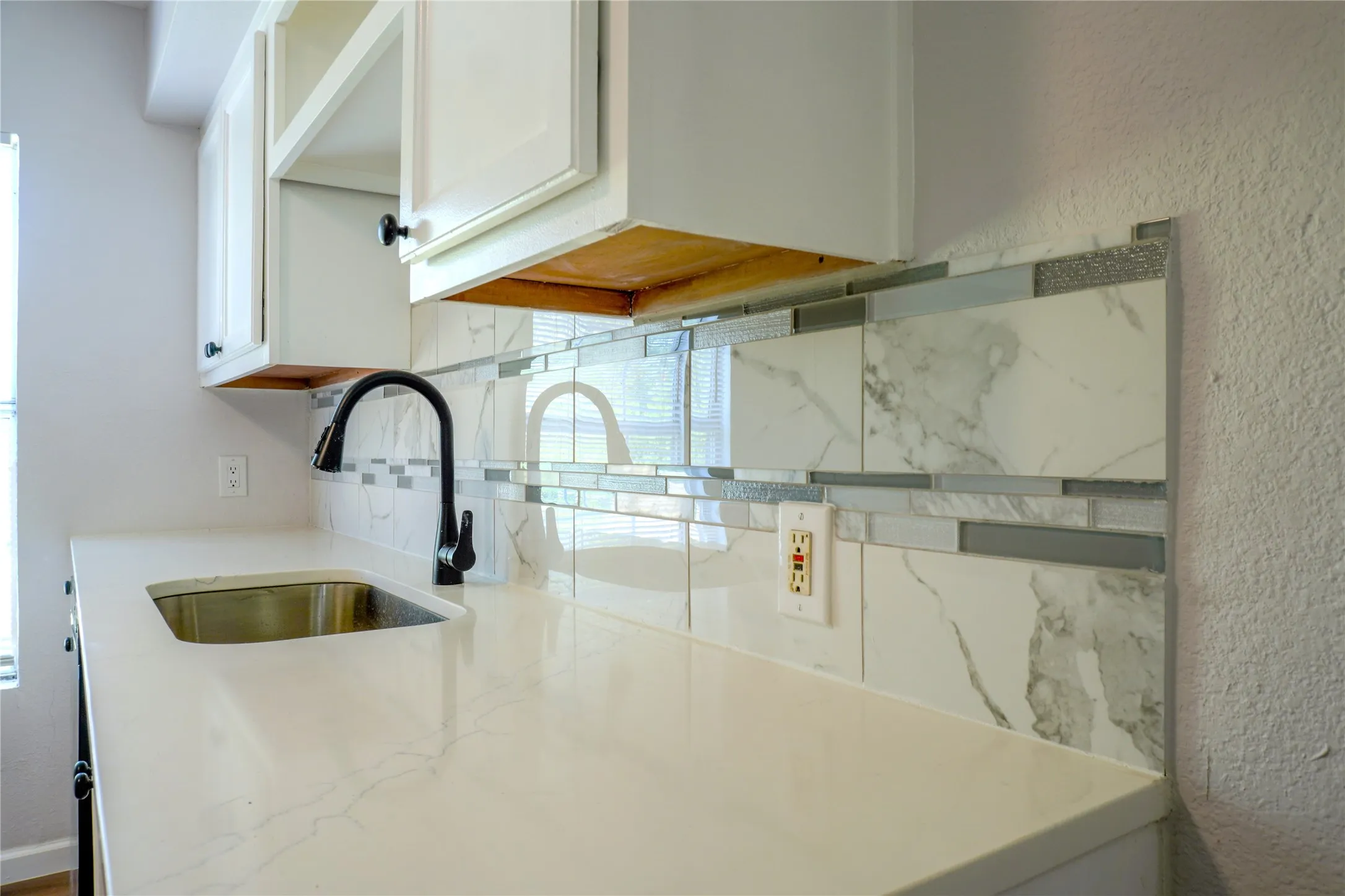 White quartz countertop, undermount stainless steel sink, matte black gooseneck faucet, and decorative tile backsplash featuring large-format stone-patterned tiles accented by glass and metallic horizontal inlay