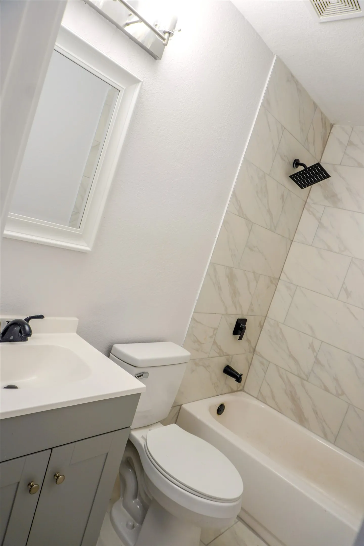 Grey vanity with a white countertop and basin, integrated matte black fixtures, and a shower-tub combination featuring tiled walls with a stone-finish pattern