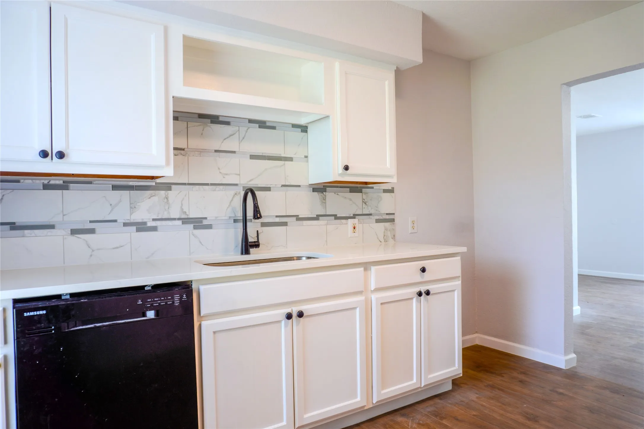 White shaker-style cabinetry, solid-surface countertop, matte black high-arch faucet, integrated dishwasher, and tiled backsplash featuring stone-patterned accents