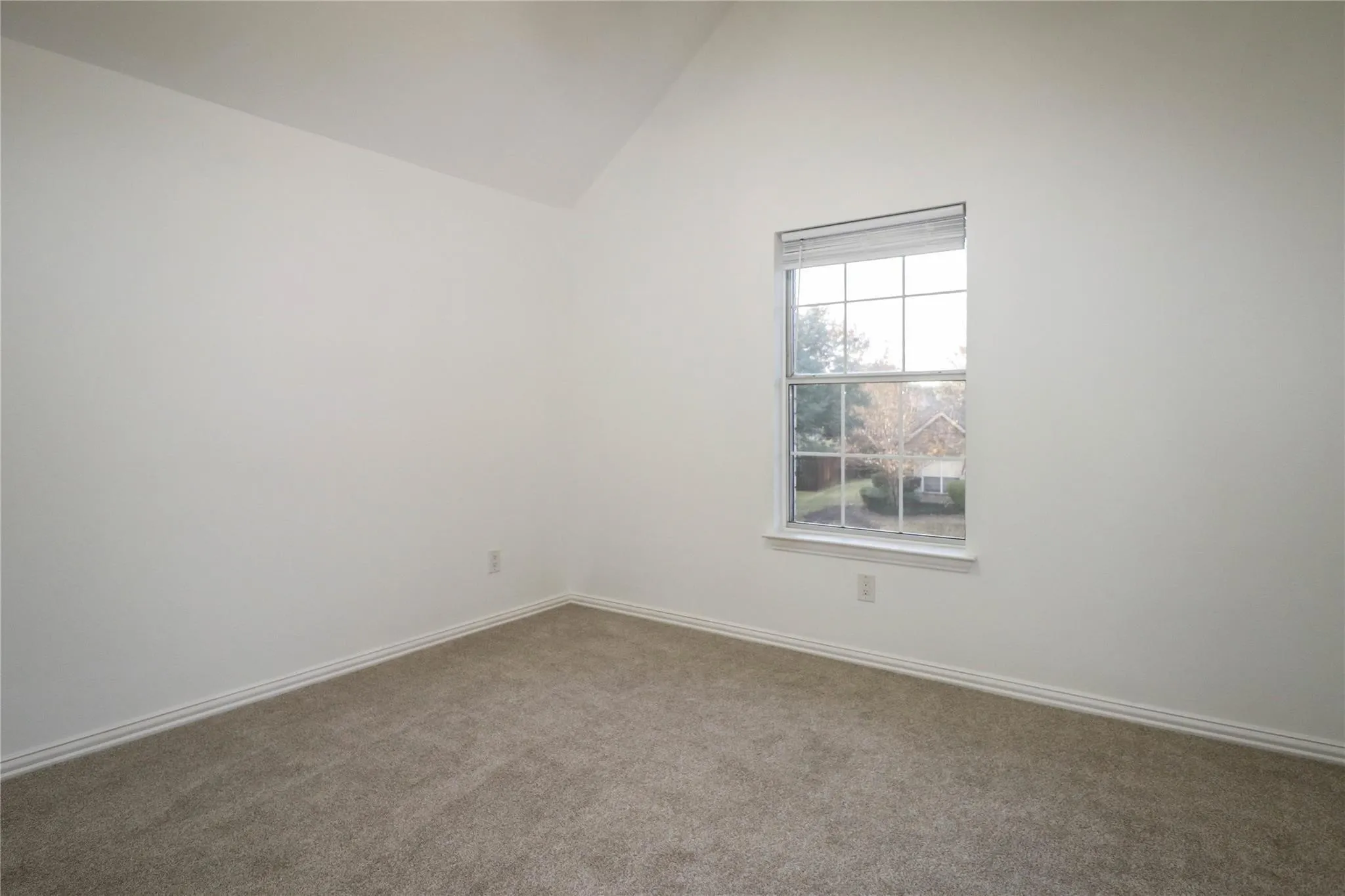Room interior featuring a vaulted ceiling, neutral wall tones, wall-to-wall carpeting, and a multi-pane window with grid detailing